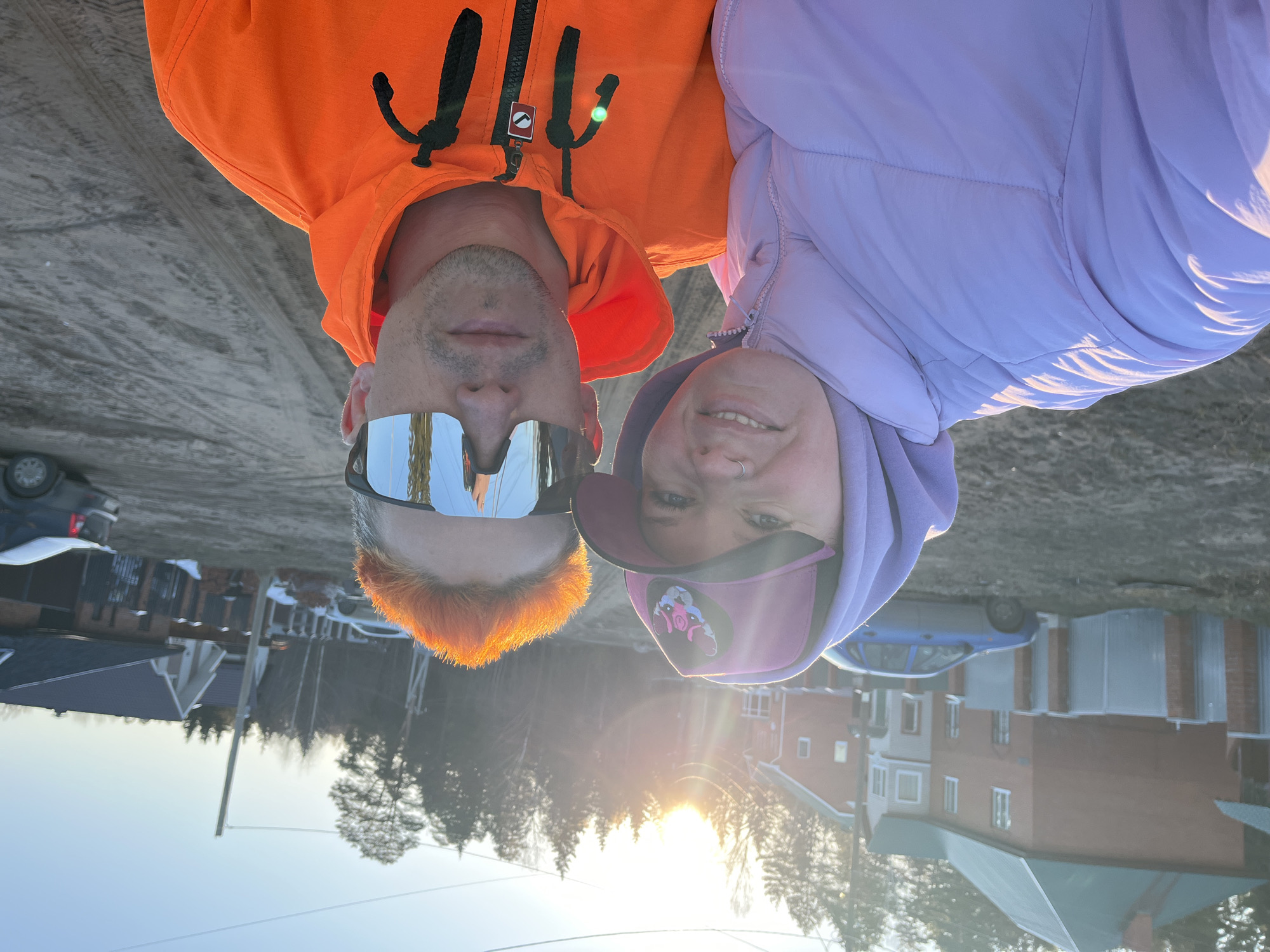 Two people are positioned close together, suggesting a selfie taken in an outdoor environment, possibly within the city of Chaikovsky, Russia, given the background architecture and ambient light. The person on the left wears an orange hooded sweatshirt with black details and mirrored sunglasses that reflect the surrounding landscape, including buildings and vehicles. The person on the right is wearing a light purple hooded jacket and a pink cap with a cartoon graphic. The background features houses and bare trees, typical of an urban setting with seasonal vegetation, and the sun, partially obscured, suggests early morning or late afternoon. The light casts long shadows on the ground.