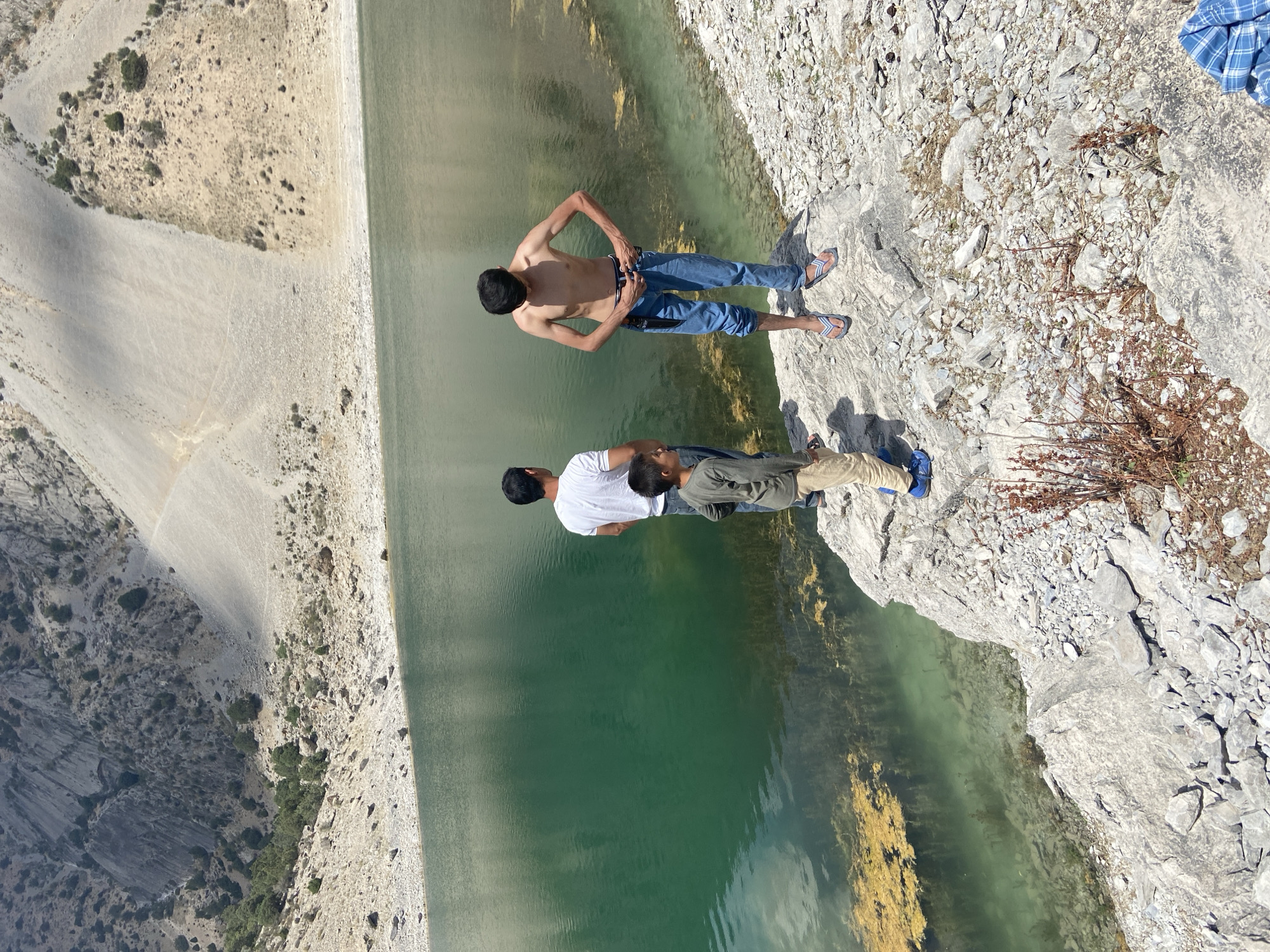 The image depicts three young men standing on the rocky edge of a body of water, possibly a reservoir or a lake, in Ghazza, Tajikistan. The scene appears to be outdoors in daylight, with clear skies suggested by the strong shadows cast on the rocky terrain. The water is a vibrant green, with patches of yellow aquatic vegetation visible beneath the surface near the shore.

The men are positioned on a light-colored, uneven rocky surface that drops off sharply to the water. One man, shirtless and wearing blue jeans and flip-flops, stands upright with his hands on his hips, looking towards the water. Another man, wearing a white t-shirt and blue jeans, stands slightly behind and to the left of the first, with his head bowed. A third man, wearing a grey long-sleeved shirt and tan pants, stands further back and to the right, facing away from the camera.

The surrounding landscape is dry and arid, characterized by rocky slopes with sparse vegetation. The terrain suggests a remote, mountainous or hilly environment. The overall impression is one of leisure and perhaps daring, given the proximity to the steep drop. There is no visible text in the image. The time of day appears to be afternoon, judging by the intensity and direction of the sunlight.