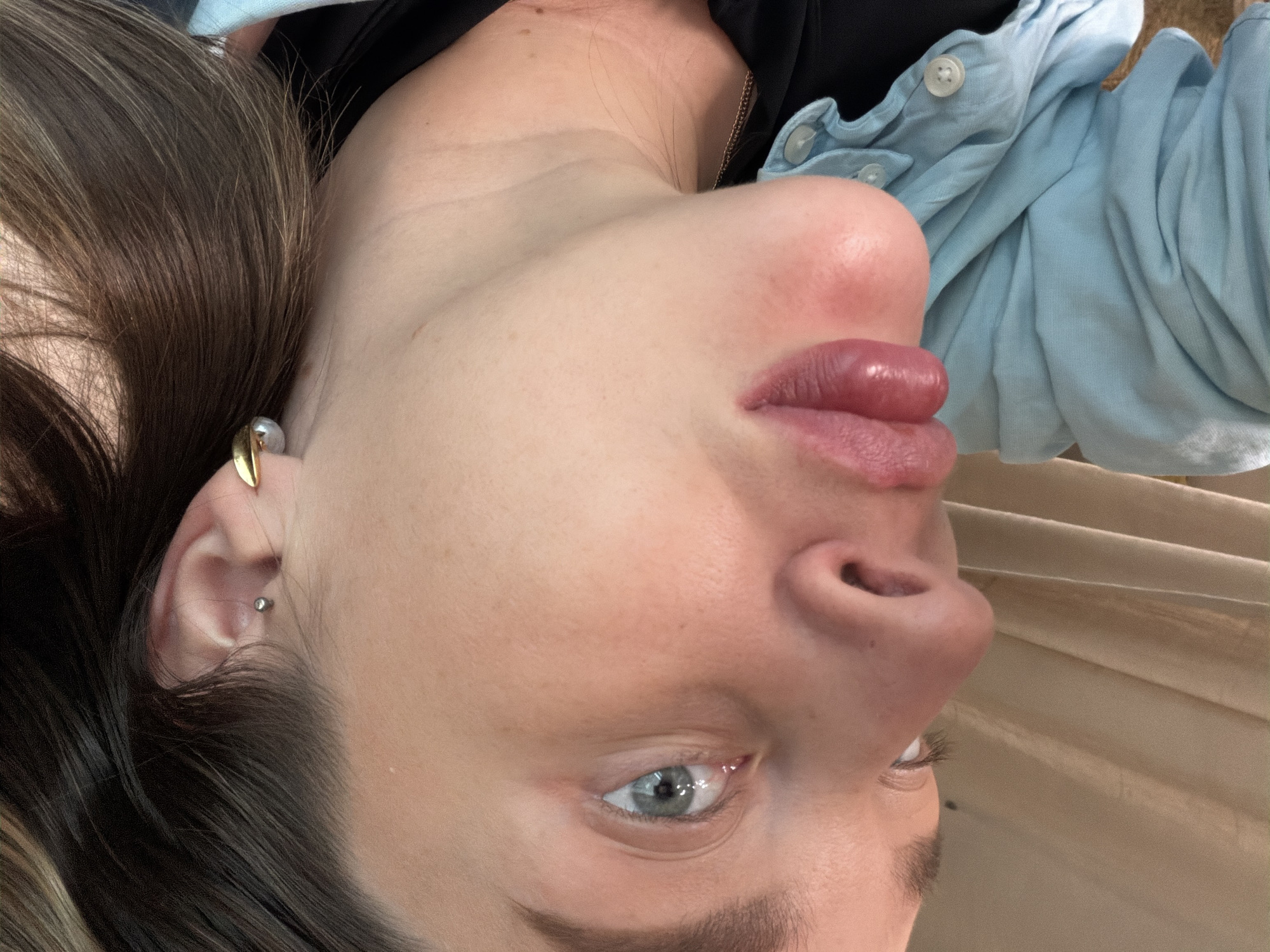A close-up, upside-down view of a person's face. The person has light skin, light brown hair, and blue eyes. They are wearing a light blue button-down shirt over a black top. They have piercings in their ear, including a stud and a gold-colored earring. The background appears to be a light brown, possibly textured surface. The location is unknown.