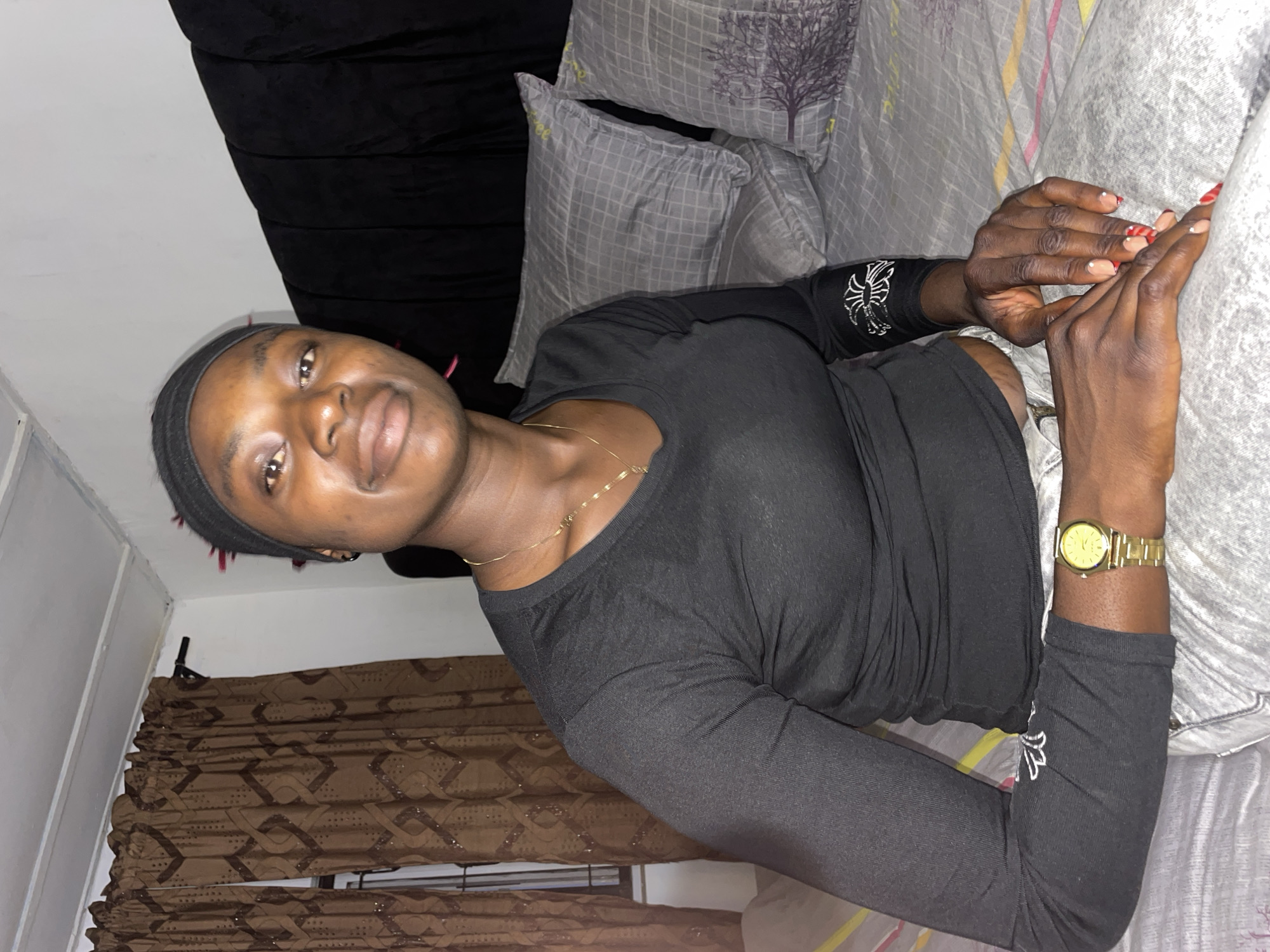 The image displays a young woman seated on a bed within an indoor residential setting, identified as being in Jos, Nigeria. The woman, with dark skin, is positioned centrally, looking directly at the camera with a slight smile. She is wearing a black long-sleeved shirt and light-colored trousers. A thin gold chain necklace with a small pendant adorns her neck, and a gold-toned wristwatch is visible on her left wrist. Her fingernails are manicured with a French tip design, incorporating red and white patterns or dots on the tips of several nails. A black headband or scarf is worn on her head, with a few reddish-pink strands of hair visible near her right temple. Her hands are clasped together, resting in front of her.
The immediate environment consists of a bed with grey patterned bedding. The pillows feature a grey grid pattern, and one pillow displays a dark tree motif. The duvet cover has horizontal yellow and pink stripes. Behind the woman is a dark, upholstered headboard. To the left side of the frame, brown curtains with a repeating pattern are drawn, partially obscuring a window that includes a visible metal security grill. The room's walls are white, and the ceiling is white with visible structural joists or paneling. The lighting suggests an artificial indoor source.