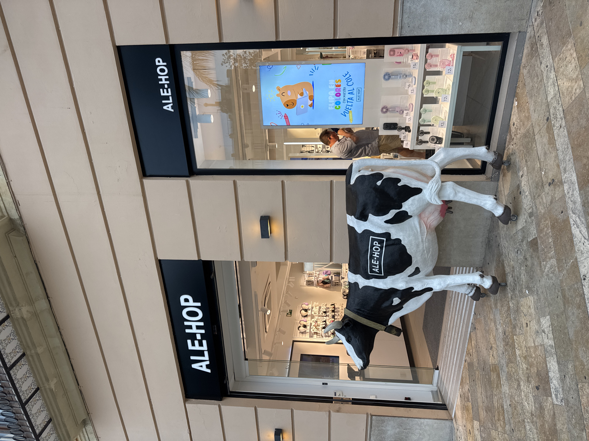 An outdoor shot captures the facade of an ALE-HOP store, a retail chain known for its trendy accessories and gadgets. The store is situated on a street in Valencia, Spain, as evidenced by the Spanish language on the signage and the architectural style. A large, black and white cow statue, a whimsical mascot for the brand, stands prominently in front of the store entrance, its presence adding a playful element to the scene. The cow statue is branded with the ALE-HOP logo. To the right of the entrance, a large display window showcases various electronic devices, including an array of colorful portable fans. A man, casually dressed, is visible inside the store, interacting with the merchandise. The store itself appears modern and well-lit, with displays of accessories and headphones visible within. The street pavement is paved with light-colored stone, characteristic of many urban areas in Spain. The overall atmosphere is that of a commercial street scene on a bright day.