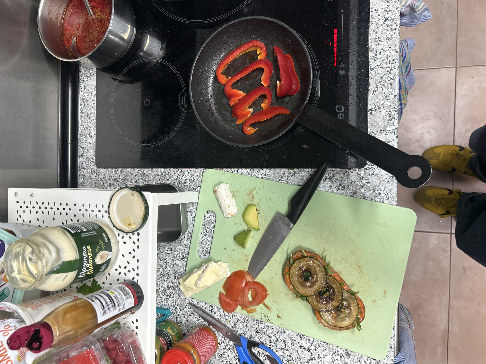 The image depicts an overhead view of a kitchen counter and floor in Málaga, Spain, with ongoing food preparation. A black induction cooktop occupies the right side of the frame, featuring a black frying pan containing several slices of red bell pepper cooking on a front burner, and a stainless steel pot filled with a red liquid, likely soup or stew, containing a silver ladle on a rear burner. The front burner under the frying pan displays an illuminated red power level indicator.
To the left of the cooktop, a light green plastic cutting board rests on a speckled granite-patterned countertop. On the cutting board, a large kitchen knife with a black handle is positioned near partially sliced red tomato pieces, two triangular avocado slices, a wedge of Brie-style cheese, and a piece of cylindrical white goat cheese or similar spread. Below these items, a prepared open-faced sandwich or bruschetta is visible, topped with three circular slices of roasted or fried zucchini, garnished with herbs. A pair of blue-handled scissors lies near a jar labeled "Baresa Pesto Genovese."
Behind the cutting board, a white perforated metal shelving unit holds a large glass jar of "Mayonesa Mahonesa Hacendado" with its lid open, a smaller white dish containing some liquid, a brown sauce bottle with a red flip-top cap, and a clear plastic container filled with red berries. A white container with a red "SMILE" logo and a percentage is also visible on the top shelf.
The floor beneath the counter is tiled with light brown square tiles. The lower portion of a person's legs and feet are visible at the bottom of the frame, wearing dark pants and yellow sneakers. A multi-colored striped kitchen towel is partially visible tucked under the countertop.