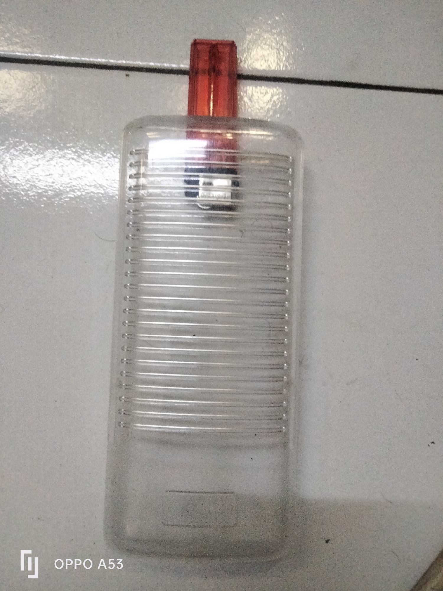 A clear plastic bottle with horizontal ridges is centered in the frame. It has a red, translucent cap. The bottle sits on a tiled surface with thin black grout lines. The overall lighting is bright, with a strong reflection coming from the left side of the bottle, suggesting a light source in that direction. The bottom left corner of the image shows the "OPPO A53" watermark, indicating the photograph was taken with an Oppo A53 smartphone. The scene does not provide enough detail to determine a specific location in Denpasar, Indonesia.