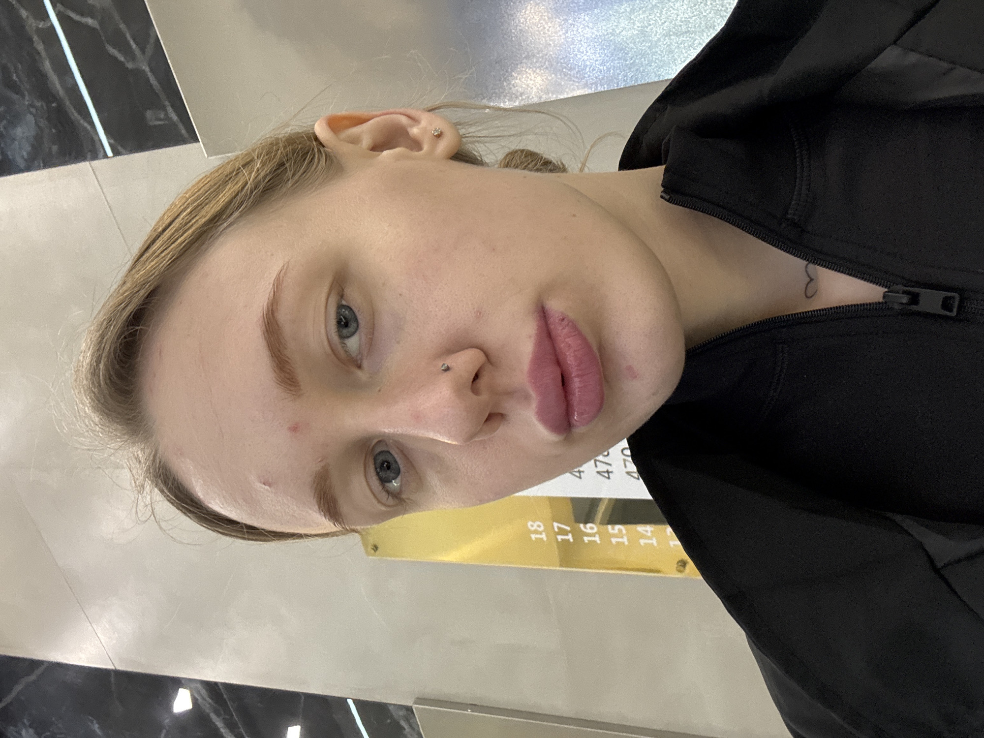 A close-up selfie of a fair-skinned person with blonde hair and blue eyes, possibly female, wearing a black zip-up jacket. The person has a nose piercing and what appears to be a small tattoo on their neck. The background includes a wall with reflective vertical strips, and a gold-colored sign with numbers, potentially indicating floor numbers (14-18) and possibly room numbers (470-476). No specific location in Rostov-on-Don is definitively discernible from the image.