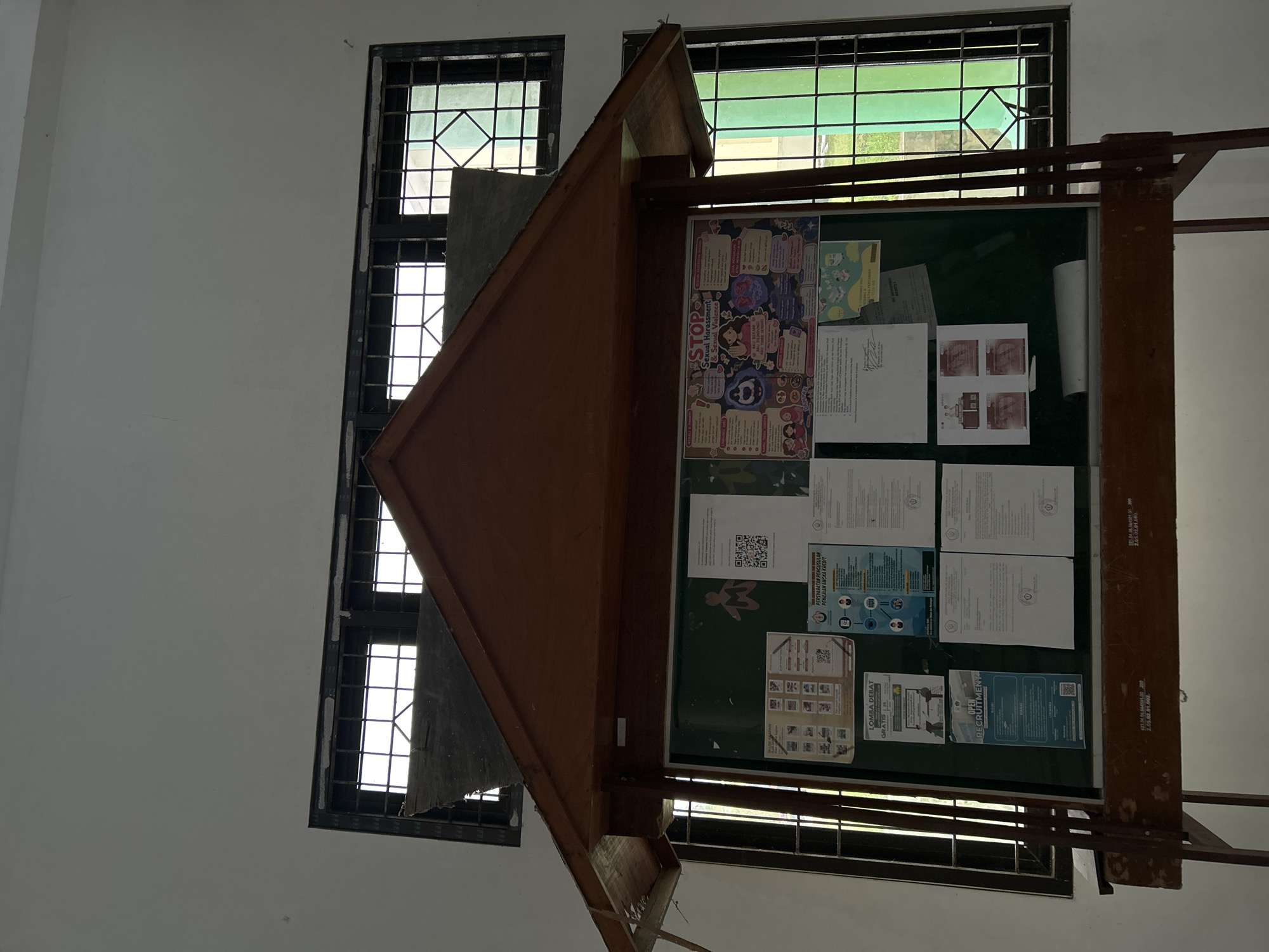 The image displays an indoor scene featuring a prominent wooden display board, likely a bulletin board, standing against a white wall. To the left and right of the board, two windows with dark frames and metal security grilles allow natural light to illuminate the space. Beyond the windows, a bright green area and some structural elements are visible, suggesting an outdoor setting with vegetation.
The display board, which appears to be a double-sided A-frame type, is covered with various printed documents, posters, and flyers, mostly in white and light colors, pinned to a dark green surface. No people are visible, indicating that no specific activity is taking place at the moment, but the board itself serves as an information hub.
Notable details include the time of day, which is daytime, evidenced by the bright light entering through the windows. The architectural style of the windows with grilles and the content of the posters suggest an institutional setting such as a school, university, or public building in Indonesia, aligning with the provided location context of Reuleut Barat.
Visible text on the posters and documents includes:
* "STOP Sexual Harassment & Sexual Violence" on a colorful poster.
* "LOMBA DEBAT" (Debate Competition) with "GRATIS 25".
* "OPEN RECRUITMENT".
* "PERSVARATAN PENGUSULAN PENILAIAN ANGKA KREDIT" (Requirements for Proposing Credit Score Assessment).
* A document reference "2.05.02.01.003.".
* Several QR codes are also present on different flyers.
The items on the board cover topics ranging from social awareness to academic or organizational recruitment and administrative procedures, highlighting its role as a communication platform.