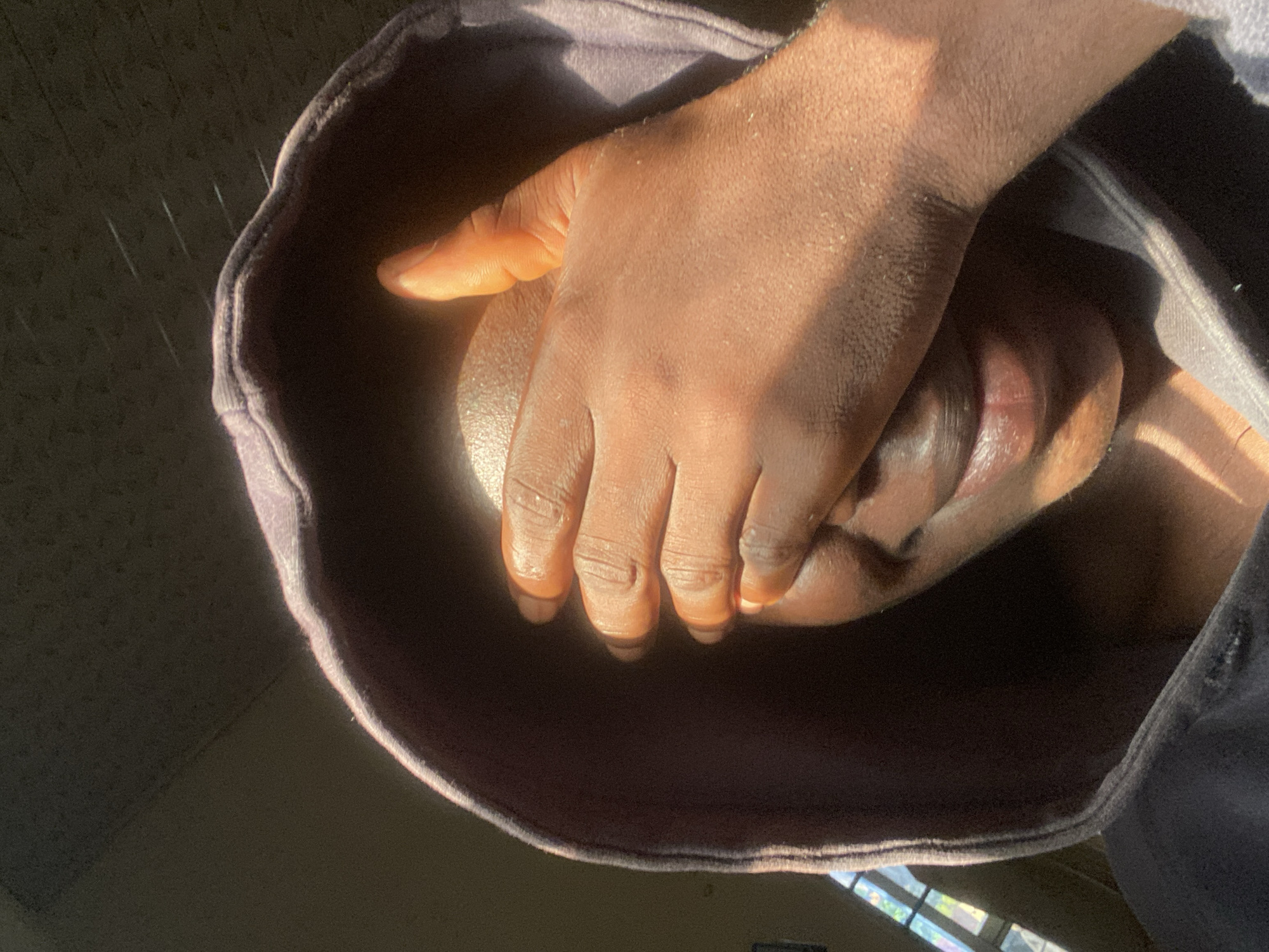In Mararaba, Nigeria, a person wearing a grey hoodie is partially obscured by their hand covering their face. Sunlight streams in, casting dramatic shadows across their skin and the fabric of the hoodie. The scene is intimate, focusing on the textures of skin and cloth under the light. The background, though out of focus, hints at a window with panes.
