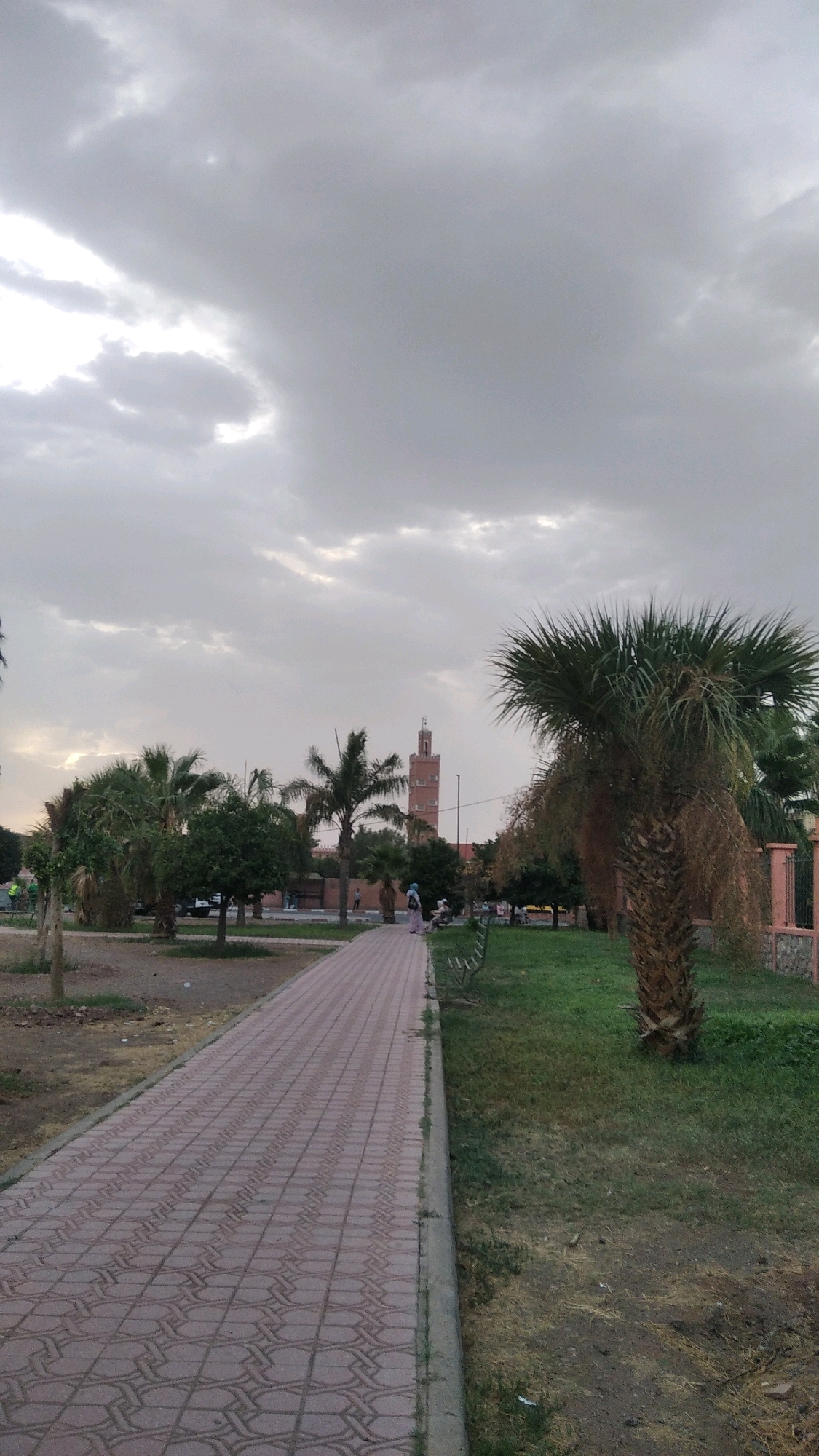 The image shows a paved walkway in a park with sparse vegetation and palm trees under a cloudy sky. A tall, slender tower with a square base and multiple levels is visible in the distance, flanked by smaller trees. Several people are walking or sitting in the park. The ground is a mixture of grass, dirt, and dry foliage. The scene appears to be in a public outdoor space, possibly a garden or square in Marrakesh, Morocco, given the architectural style of the tower and the presence of palm trees.