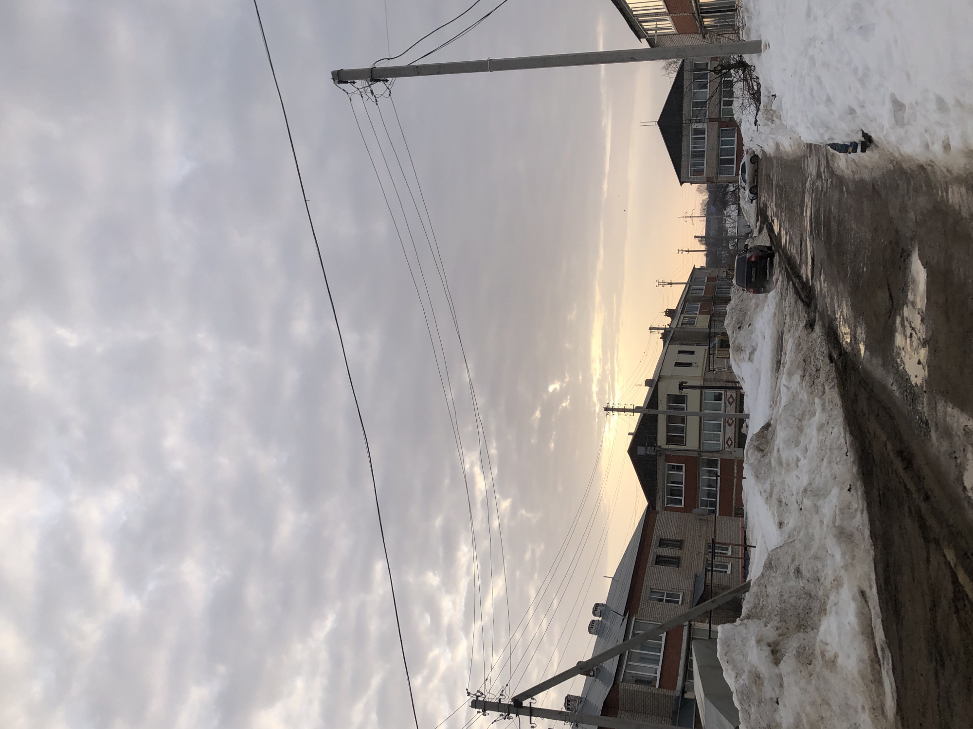 This image depicts a residential street in the city of Фоки, Russia, characterized by conditions typical of a winter-to-spring transition. The sky is largely overcast with stratocumulus clouds, exhibiting a distinct low-lying golden-orange glow on the horizon, indicative of either sunrise or sunset.
The unpaved or poorly maintained street surface is muddy, wet, and contains numerous water-filled potholes and prominent vehicle tire tracks. Large, dirty snowdrifts line both sides of the road, exhibiting significant melting and revealing patches of dark, exposed mud.
Several multi-story residential buildings flank the street. On the immediate left, a two-story building constructed of light-colored brick with red-orange accents features white-framed windows and a dark, sloped roof with multiple chimneys. Further down the street, additional residential structures with varying facades and window configurations are visible on both sides.
Two concrete utility poles are present; one on the left foreground and another on the right midground, both supporting multiple overhead power lines that extend across the scene.
A dark-colored sedan is positioned on the left side of the muddy road, facing away from the viewer. A lighter-colored sedan is partially visible further down the road on the right. No individuals, specific actions, or direct interactions are observable within the frame.