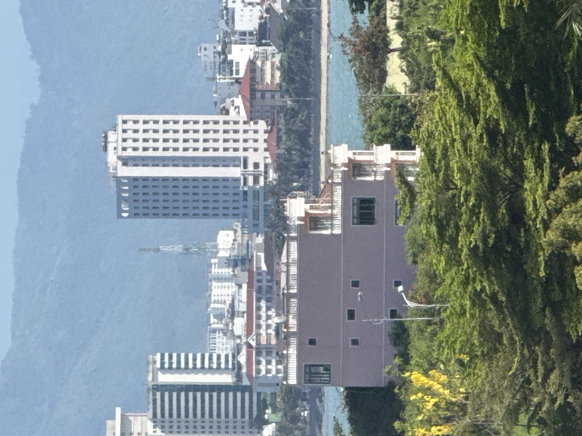 The image is a distant, slightly blurry view of a cityscape with a body of water and mountains in the background. In the foreground, there is lush green foliage, including trees with bright yellow flowers. Behind the foliage, a light pink building with multiple windows is visible. Further in the background, there are various other buildings, including tall, modern high-rises with many windows, and some lower-rise buildings with red roofs. A tall communication tower stands between two of the high-rises. The body of water appears to be the sea or a large bay, with a faint, possibly a small boat, visible on it. The mountains in the far distance are hazy and blue. The scene suggests a coastal city in Vietnam, likely Bac Nha Trang Ward, known for its beaches and mountainous backdrop. There are no people or discernible actions depicted in the image.