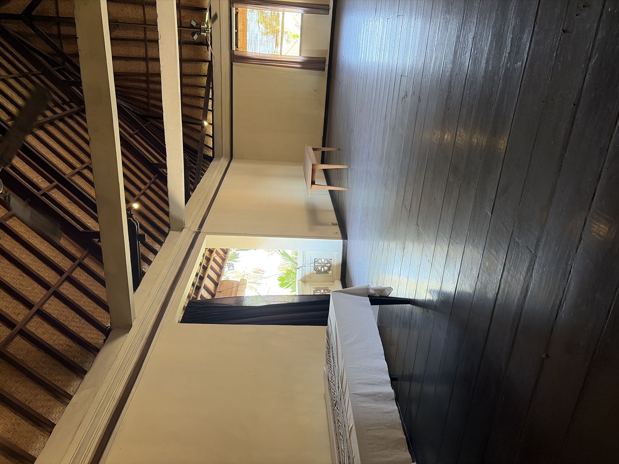 This image depicts an interior scene within a building, likely a room in Ubud District, Indonesia, characterized by traditional architectural elements. The room features a high, thatched roof with exposed wooden beams and rafters, suggesting a rustic or tropical aesthetic. Natural light streams in through open windows and is augmented by ceiling lights.
The walls are painted a pale yellow, contrasting with the dark, polished wooden floorboards. In the foreground, a rectangular table is covered with a white tablecloth adorned with a black and white patterned runner. The room appears spacious and sparsely furnished.
Further into the room, a small, light-colored wooden table with four legs stands against the wall. Two windows are visible: one on the left with dark curtains partially drawn, revealing glimpses of lush greenery outside, and another on the right with bamboo blinds that allow sunlight to filter through, illuminating the floor. Two ceiling fans are mounted on the beams, indicating a warm climate. The overall impression is one of a tranquil, airy space with a connection to nature.