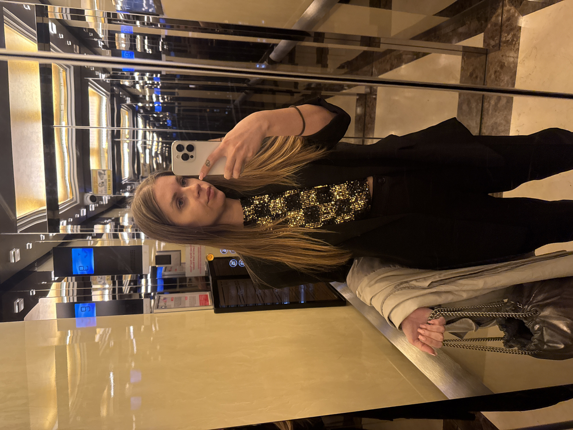 A woman with long, light brown hair and fair skin is taking a selfie in an elevator with mirrored walls. She is holding a white smartphone in her left hand, wearing a black top with a shimmering, checkered pattern, and a black jacket. She also has a black bracelet on her left wrist. She is standing next to a silver handbag with chain straps and a light-colored jacket or coat. The elevator's mirrored interior reflects the elevator buttons and display indicating floor 8. The location is suggested to be in Batumi, Georgia. The flooring of the building or elevator can be seen in the reflection, it is a light cream color with brown veining.