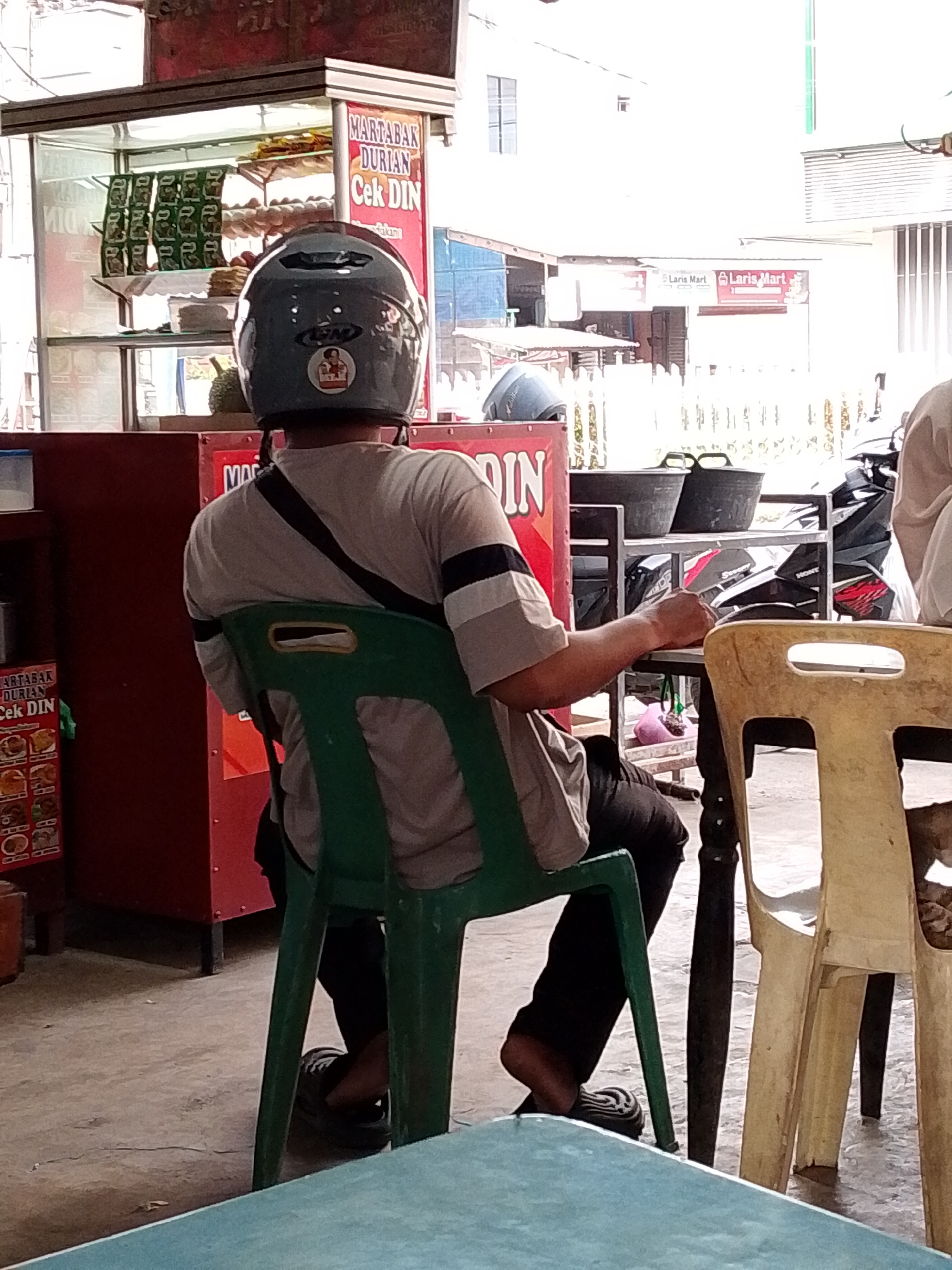A person wearing a helmet sits in a green plastic chair facing away from the camera at an outdoor food stall in Lhokseumawe, Indonesia. The stall has a red cabinet with a sign reading "MARTABAK DURIAN Cek DIN" and displays various food items. The person is wearing a grey shirt with black and white stripes on the sleeves and dark pants. Their left hand is holding a piece of paper or a small card. In the background, other food stalls and motorcycles are visible, along with signage for "Laris Mart." The ground is paved with concrete.