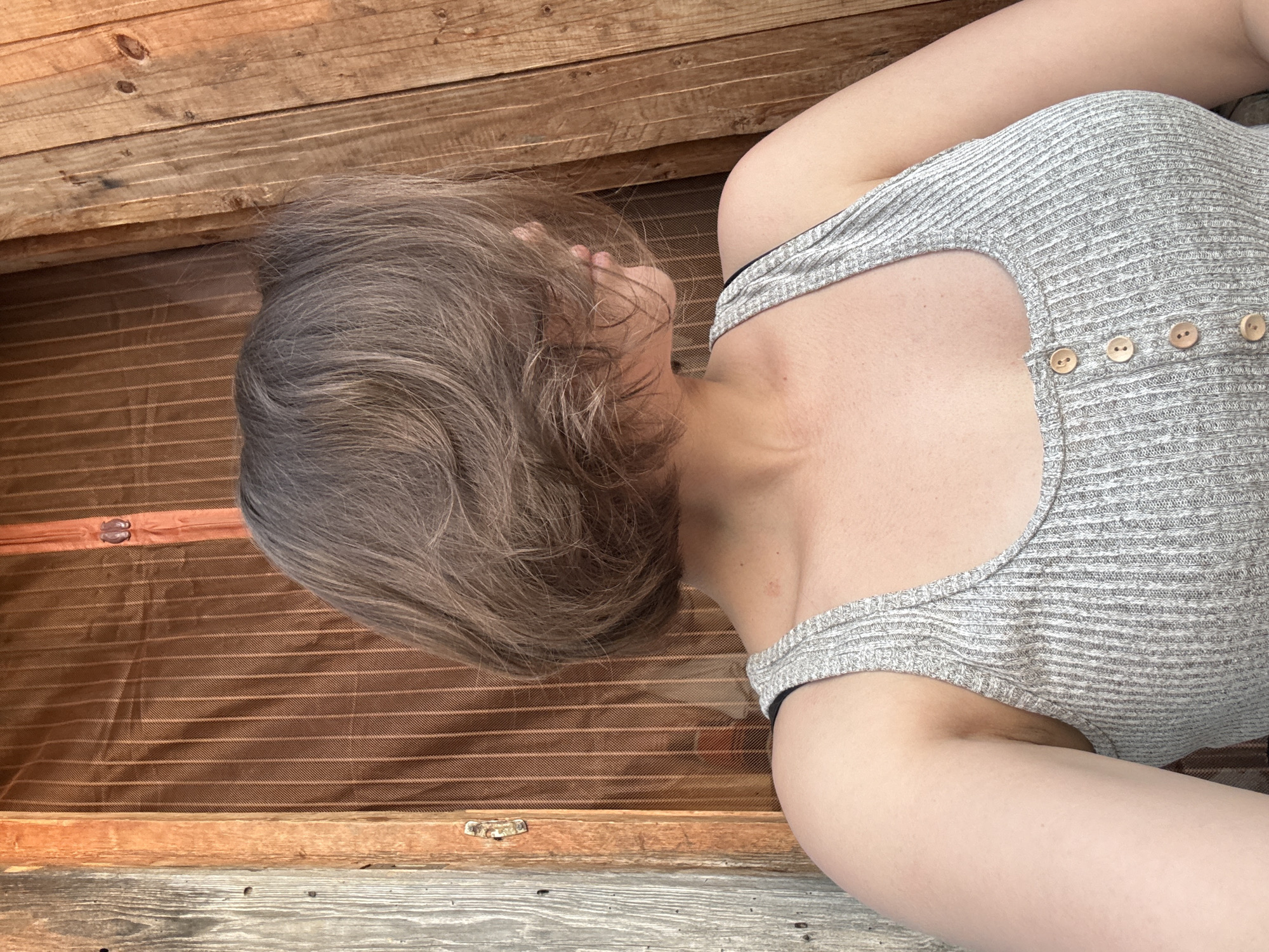 The image shows a person lying inside a wooden trunk. The trunk is lined with brown fabric with light-colored stripes. The person has short, gray-brown hair and is wearing a gray, ribbed tank top with wooden buttons on the side. The wooden planks of the trunk's exterior are visible. There is no indication that the location is in Lenino, Russia.