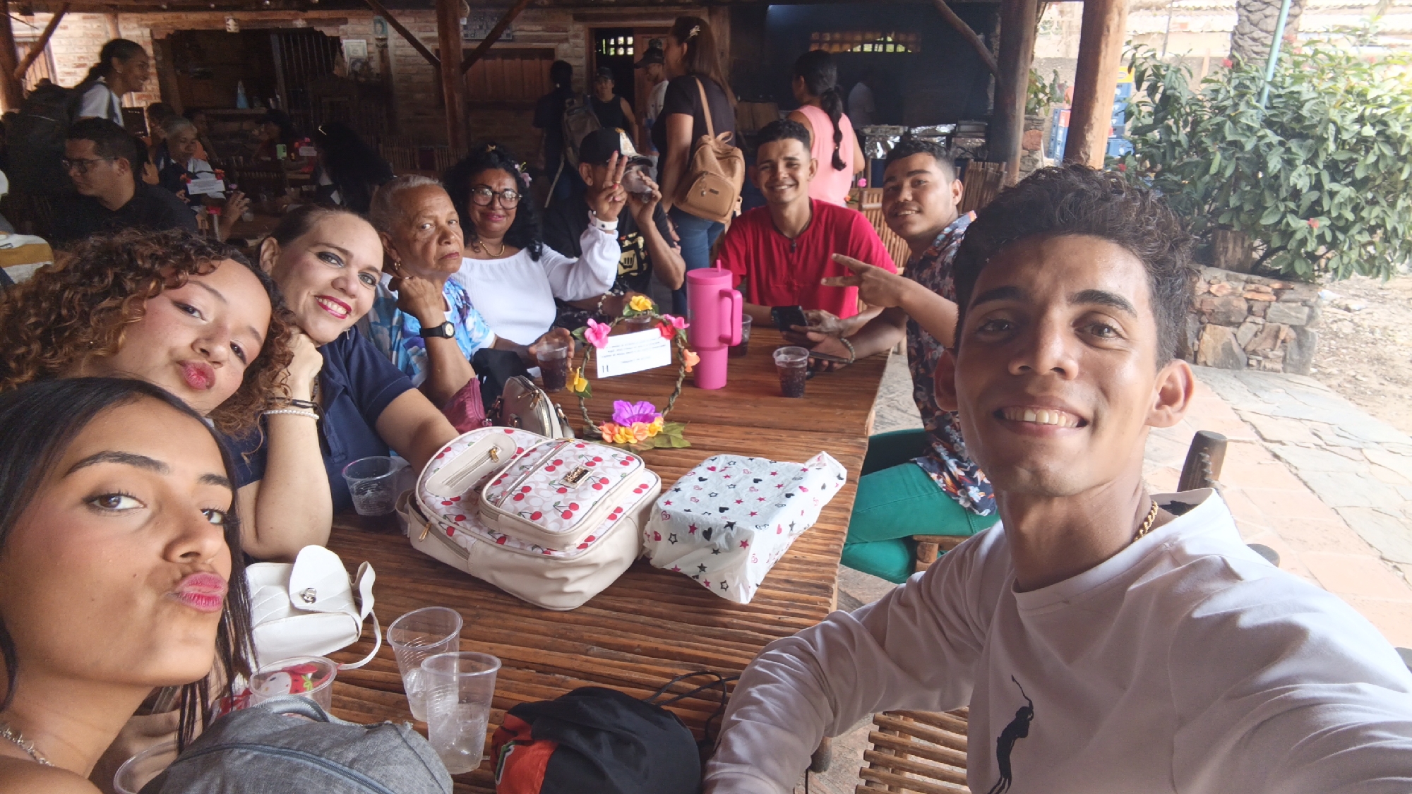 The image shows a group of approximately 12 people seated at a long wooden table. They are smiling and posing for a selfie taken by a young man in a white long-sleeved shirt. The table is covered with bags, cups, a pink thermos, and a decorative floral wreath. Some people in the background are moving around. The location appears to be an open-air restaurant or patio with a thatched roof and stone walls, possibly in Juan Griego, Venezuela, based on the appearance of the vegetation and building materials. The weather looks sunny.