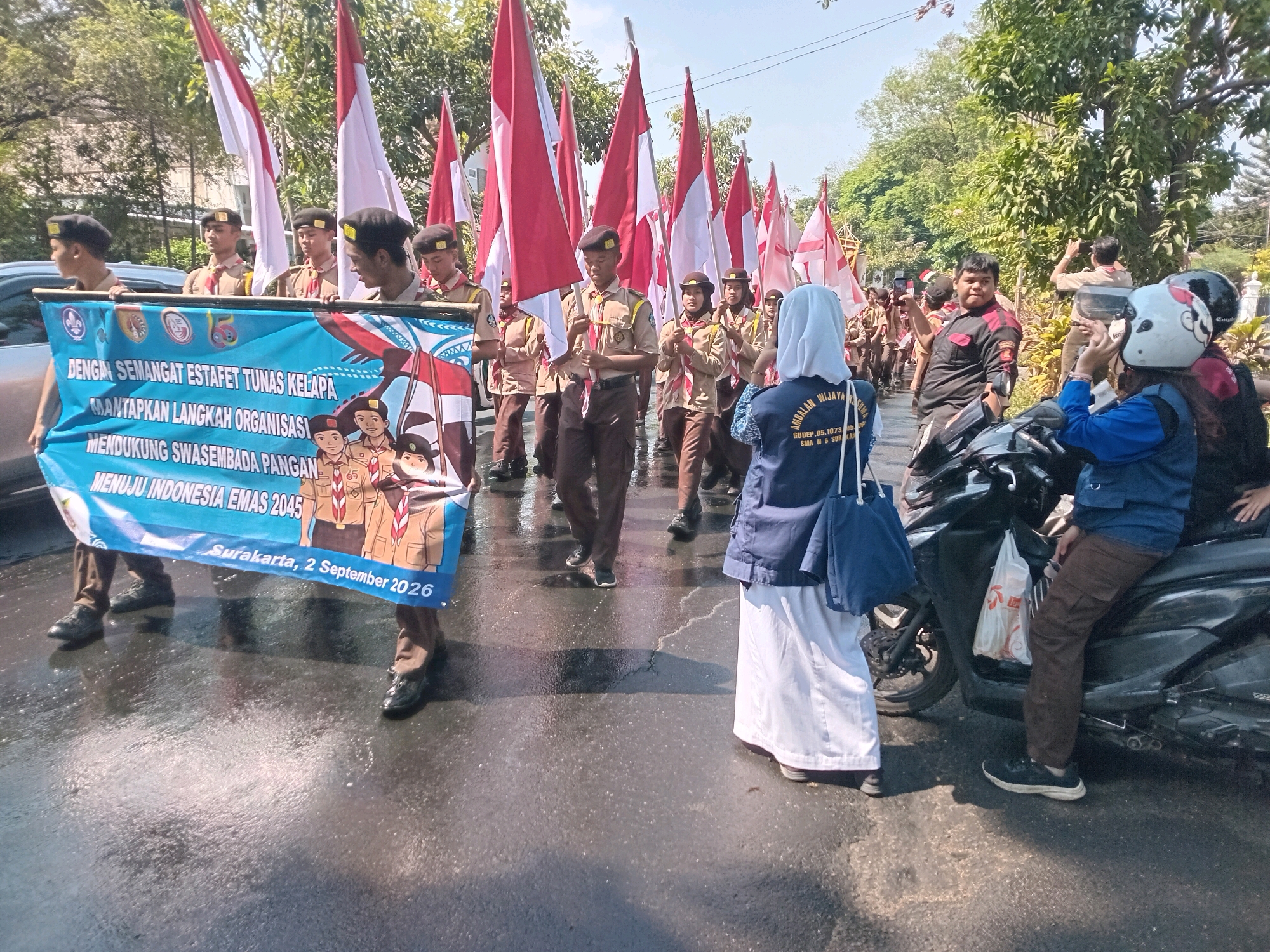 A group of young people, likely scouts, are participating in a parade in Surakarta, Indonesia. They are carrying a large blue banner with text in Indonesian that reads "DENGAN SEMANGAT ESTAFET TUNAS KELAPA MANTAPKAN LANGKAH ORGANISASI MENDUKUNG SWASEMBADA PANGAN MENUJU INDONESIA EMAS 2045 Surakarta, 2 September 2026". The banner depicts illustrations of scouts. Many of the participants are wearing brown uniforms and berets, while others are in casual clothing. They are walking on a wet asphalt road, possibly due to recent rain, as evidenced by reflections on the surface. Several Indonesian flags, red and white, are being carried by individuals in the parade. In the background, trees and buildings are visible. Some people on motorcycles are present at the side of the road, observing the parade. One person on a motorcycle is wearing a helmet and looking at their phone. Another person, wearing a white hijab and a dark blue jacket, is taking a photo with their phone. The weather appears to be sunny.