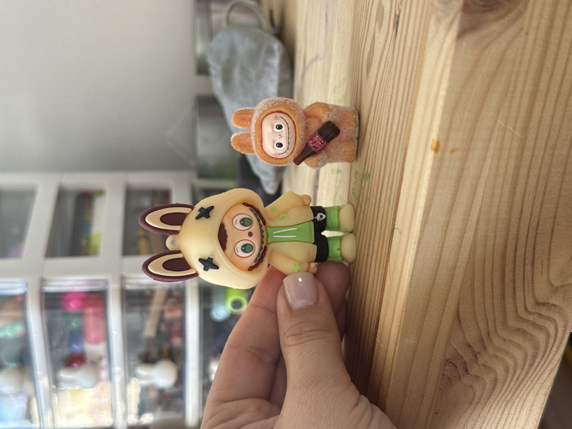 In the city of Lyubertsy, Russia, on a wooden surface, two collectible figures are displayed. The figure on the left, held by a hand with a light pink nail polish, is a character wearing a yellow hoodie with rabbit ears, green shorts, and green shoes. The figure on the right is a smaller, fuzzy orange character wearing a matching hoodie and holding a miniature Coca-Cola bottle. In the background, a multi-drawer storage unit filled with various small items and toys is visible. The scene appears to be an indoor setting, possibly a home or a studio, with diffused lighting.