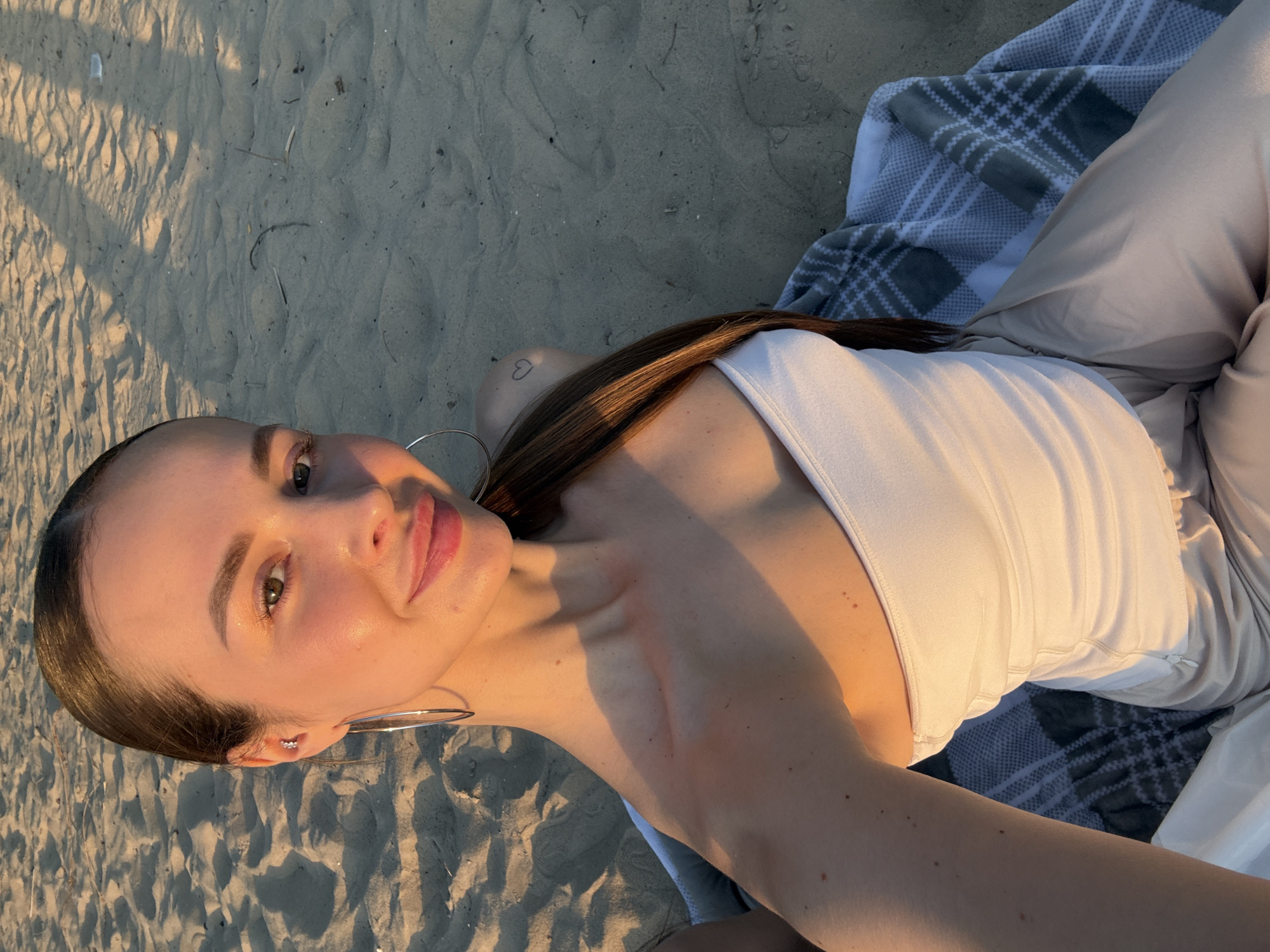 A young woman lies on her back on a sandy beach, possibly near a riverbank, as there's no evidence of an ocean. She wears a white tank top and light-colored pants, partially obscured by a gray and white plaid blanket underneath her. The woman has long brown hair, silver hoop earrings, and a small heart tattoo on her upper chest. The late afternoon sun casts a warm glow and distinct shadows across her face and the sand. The location is indeterminate without additional landmarks or context. The city of Kyiv cannot be confirmed.