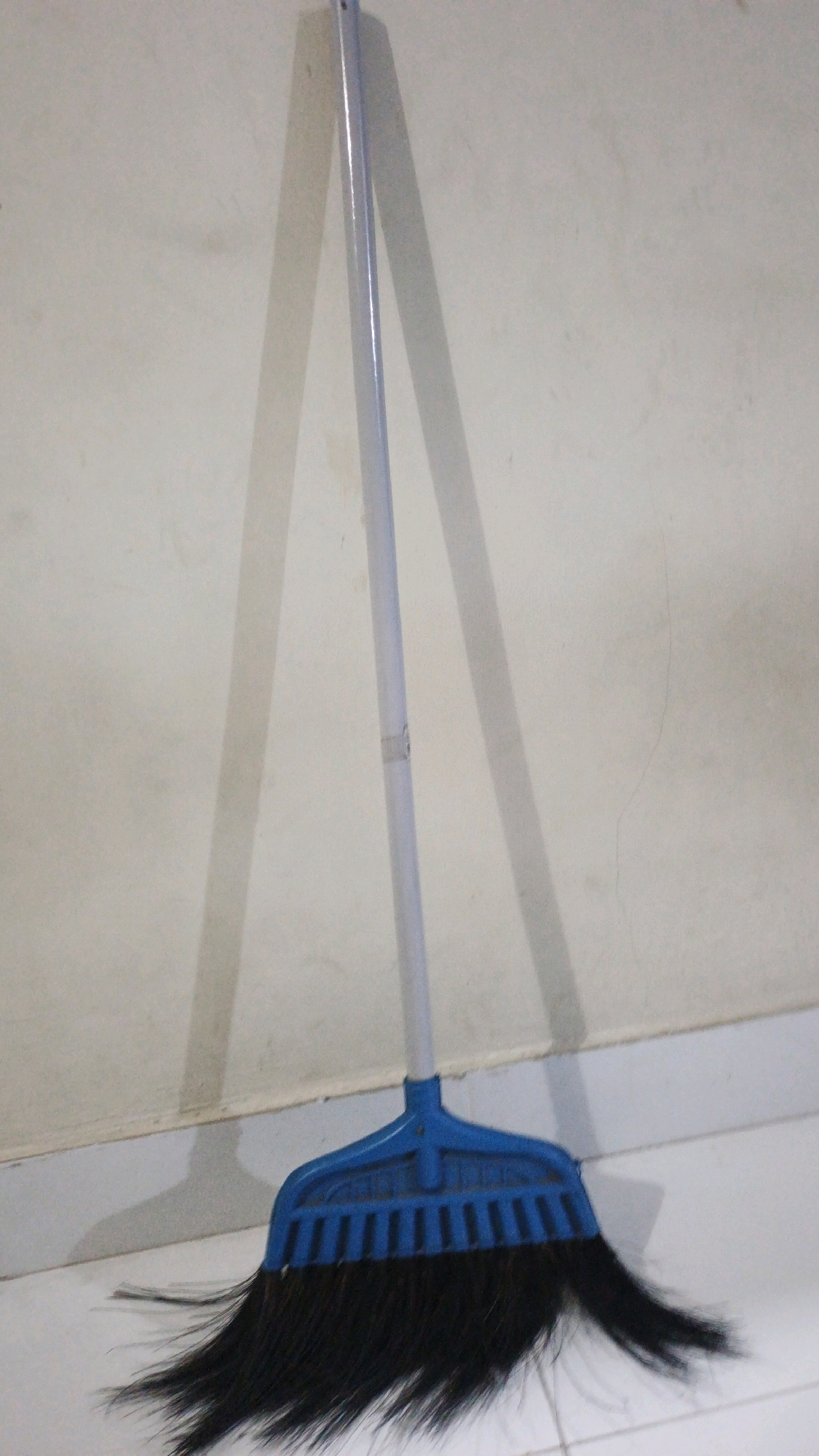 A broom with a blue plastic head and black bristles leans against a light-colored wall. The broom has a long, light-colored handle. Its shadow is cast on the wall behind it. The broom is positioned on a tiled floor. The scene appears to be indoors, possibly in a residential or commercial space in Denpasar, Indonesia. There are no people visible in the image.