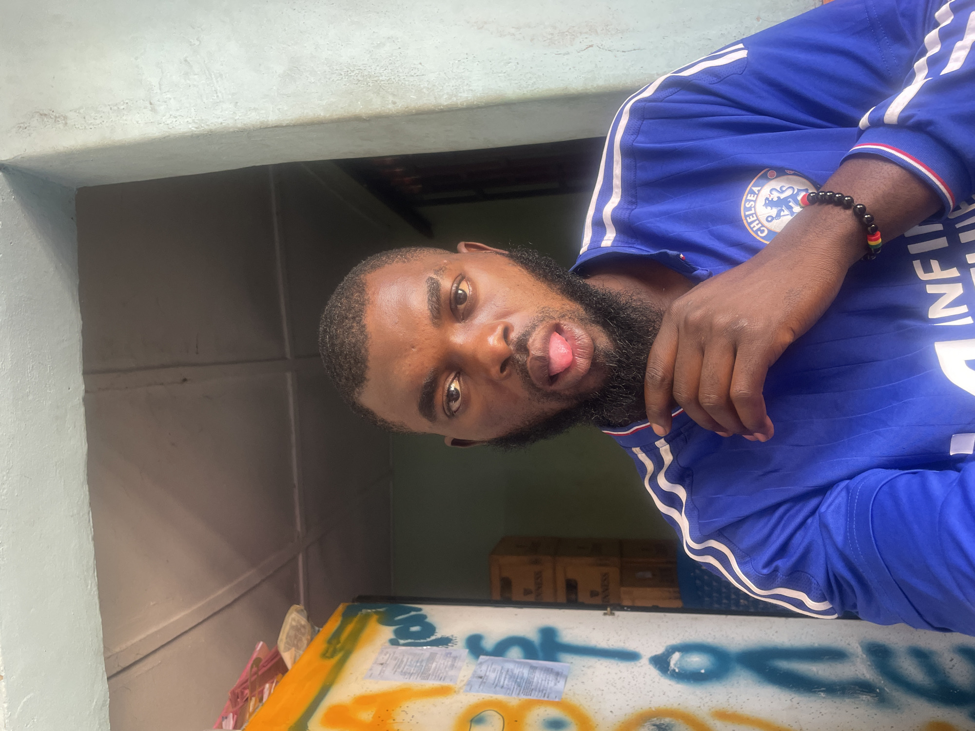 A man with a beard sticks out his tongue, posing for a selfie indoors. He is wearing a blue Chelsea F.C. jersey and a beaded bracelet. Behind him, on the left, is a wall covered in graffiti with yellow and blue spray paint. To the right, there are stacks of boxes, possibly crates of drinks. The ceiling appears to be made of wood panels. The background suggests an indoor or semi-covered area, potentially a shop or storage space. The lighting is natural and seems to be coming from the front. While the image depicts a common scene, the specific location in Jos, Nigeria, cannot be determined from the visual information alone.