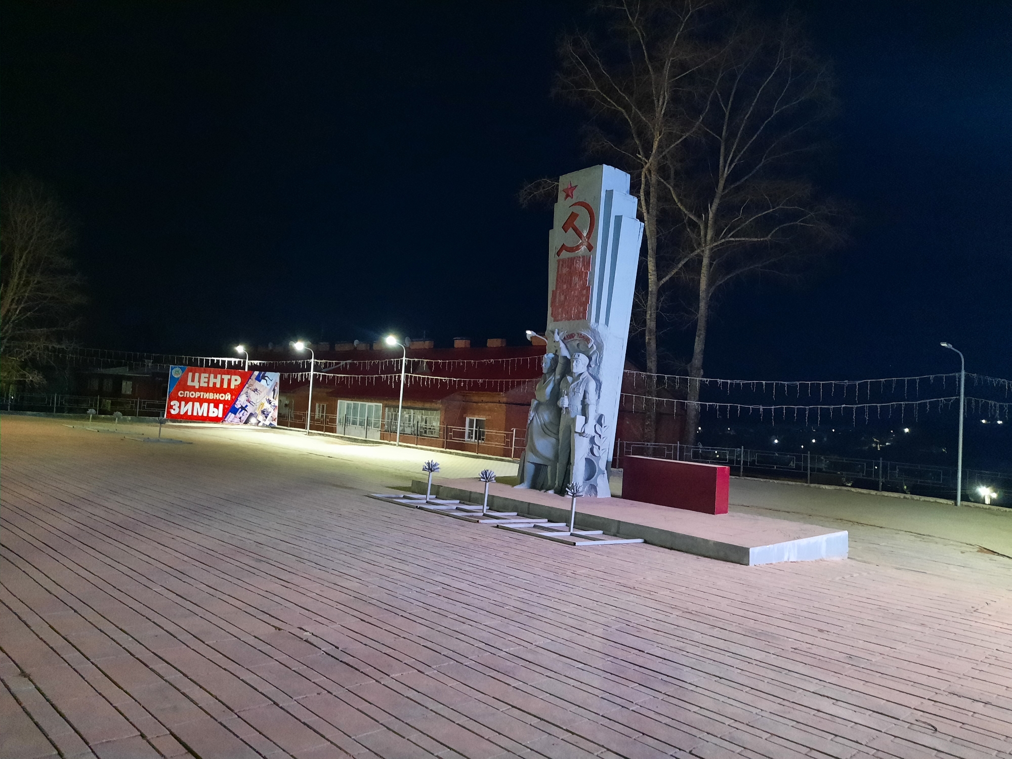 The image shows a monument in Krasnogorskoye, Russia, at night. The monument features a stylized depiction of soldiers and the communist hammer and sickle symbol. The ground is paved with rectangular blocks. A sign in Russian advertises a winter sports center. String lights are visible in the background. Several saplings stand in front of the monument. A small building with a red roof is also visible in the background. The sky is dark.