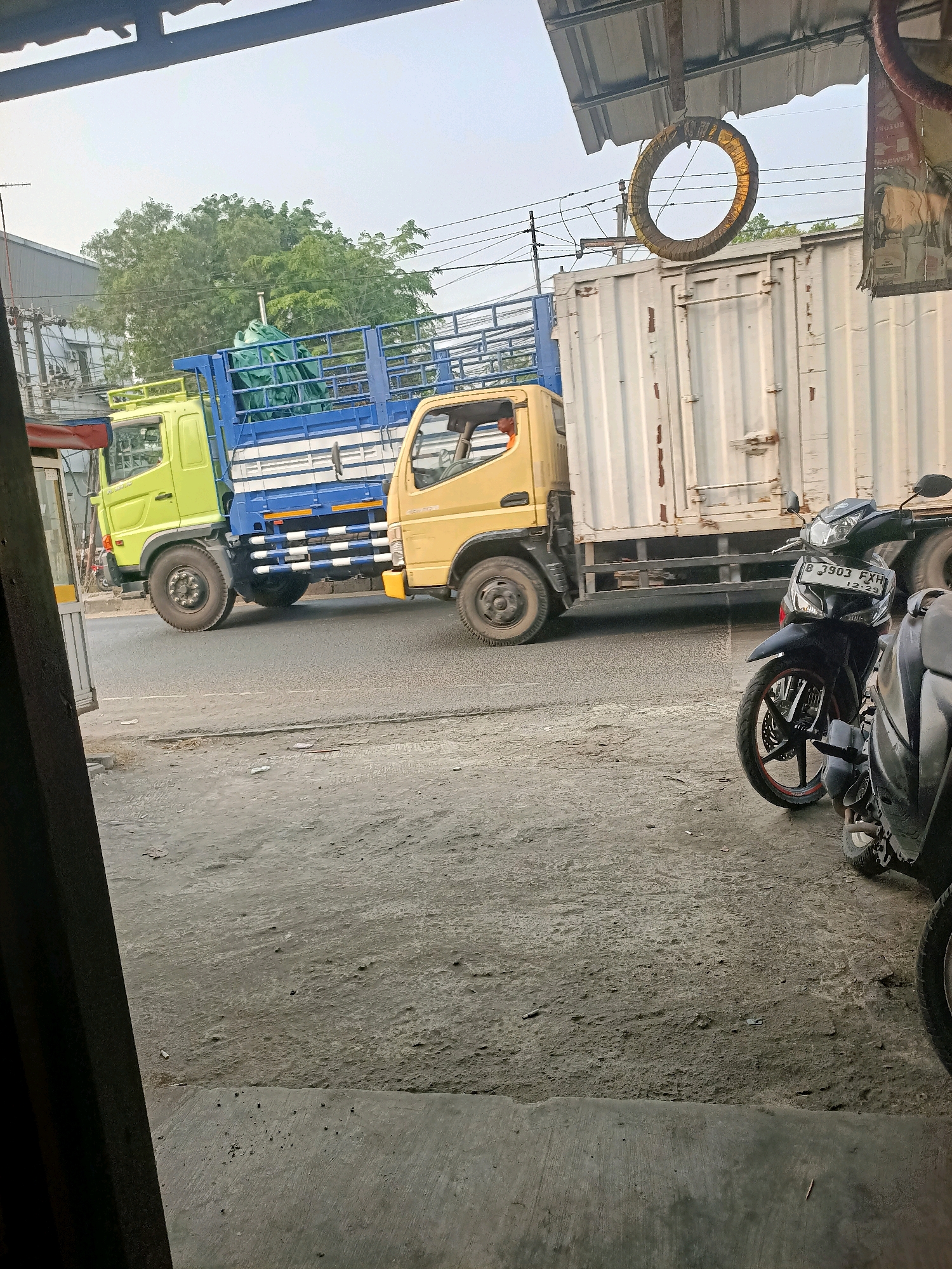 In Gandasari, Indonesia, a light yellow truck with a blue cage bed, partially covered with a green tarp, is parked on a street next to a white container truck. The truck bed is lined with blue and white striped planks. The scene also includes two parked motorcycles, one black with red accents and a license plate reading "B 3903 FXH". A circular object, wrapped in yellow material, hangs from an overhead structure. The ground is a mixture of dirt and concrete, with scattered debris. The overall environment suggests an urban or semi-urban setting.