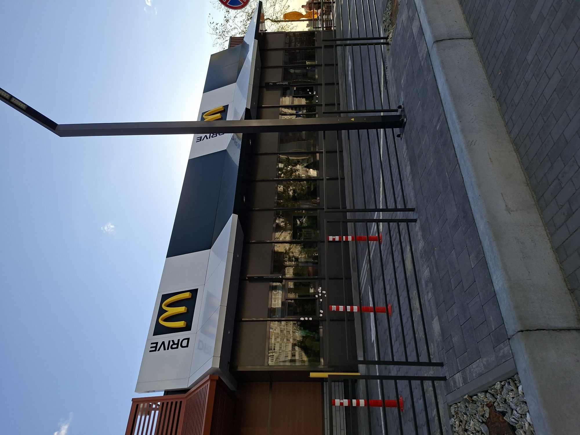 The image shows a McDonald's restaurant with a drive-thru lane. The building is modern with large glass windows. The exterior is dark gray and white, with the iconic golden arches of McDonald's prominently displayed. The drive-thru lane is marked with black metal barriers and red and white striped bollards. A tall black light pole stands in front of the building. The ground is paved with dark gray interlocking bricks, and there is a concrete curb. In the background, trees and a building with multiple windows are visible. The sky is clear blue with a few wispy clouds.