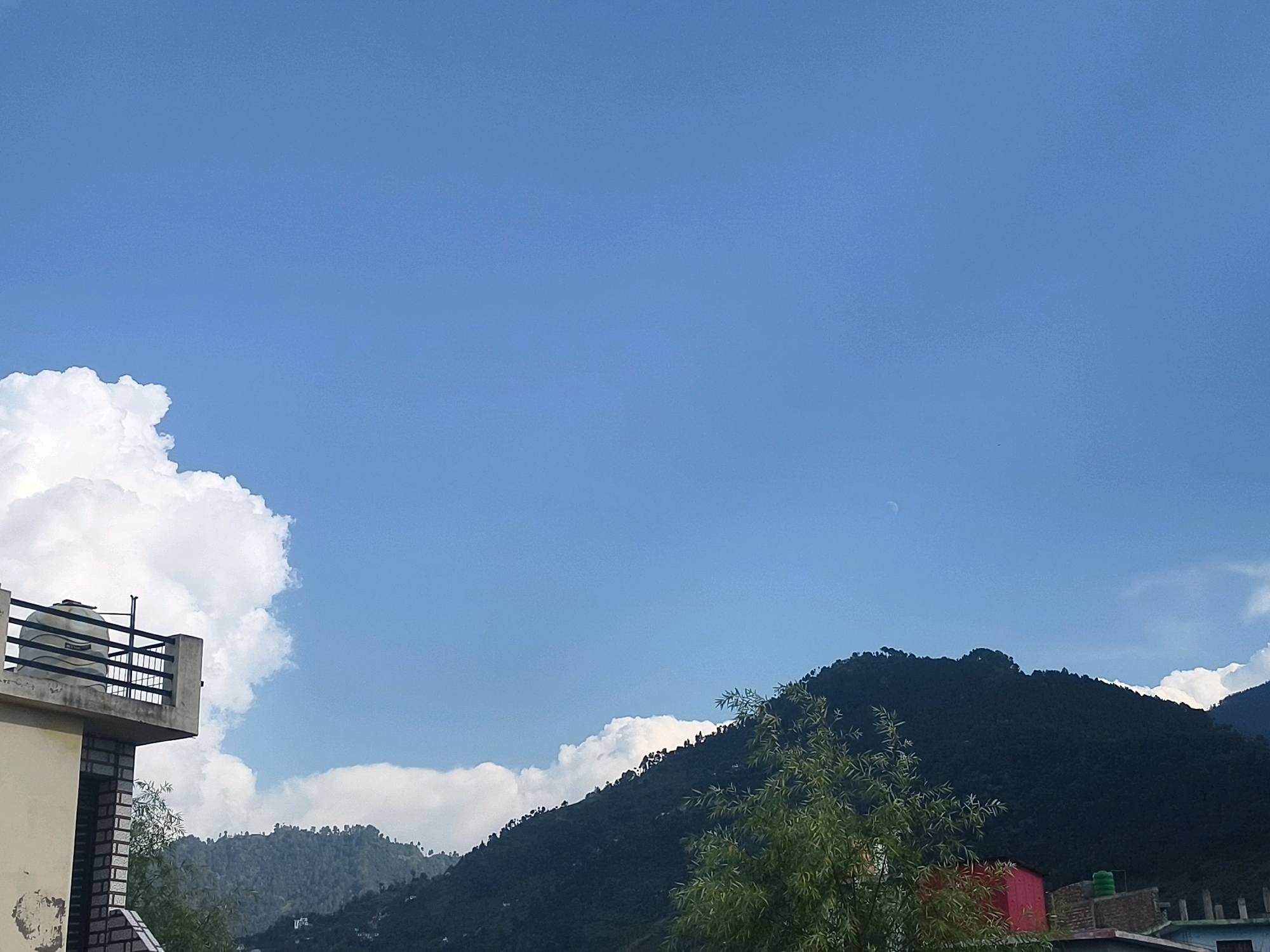 The image captures a serene natural landscape with a clear blue sky. In the foreground, a portion of a building is visible on the left, featuring a balcony with a railing and a water tank. The building's facade is a light yellow with some brickwork near a window. Beyond the building, a lush green mountain range dominates the scene, its slopes covered in dense foliage. A few white clouds are scattered across the sky, and a faint crescent moon is visible near the top of the mountain. In the lower right corner, parts of other buildings, including a red structure and a water tank, are visible, suggesting a residential area nestled within the mountainous terrain. The overall scene depicts a peaceful, possibly rural or semi-urban, environment in the city of Poonch, India, characterized by its natural beauty and the coexistence of human settlements with the mountains.