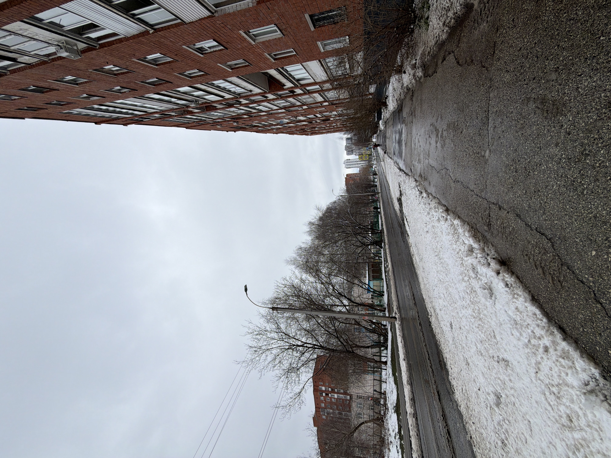 An eye-level, slightly off-kilter outdoor shot depicts a street in Izhevsk, Russia. A tall, red-brick apartment building looms on the left. The street is paved, with patches of melting snow lining the edges. Bare trees stand between the road and another brick building, partially visible in the distance, further down the street. A street lamp stands prominently on the left side of the road. The sky is overcast and grey.