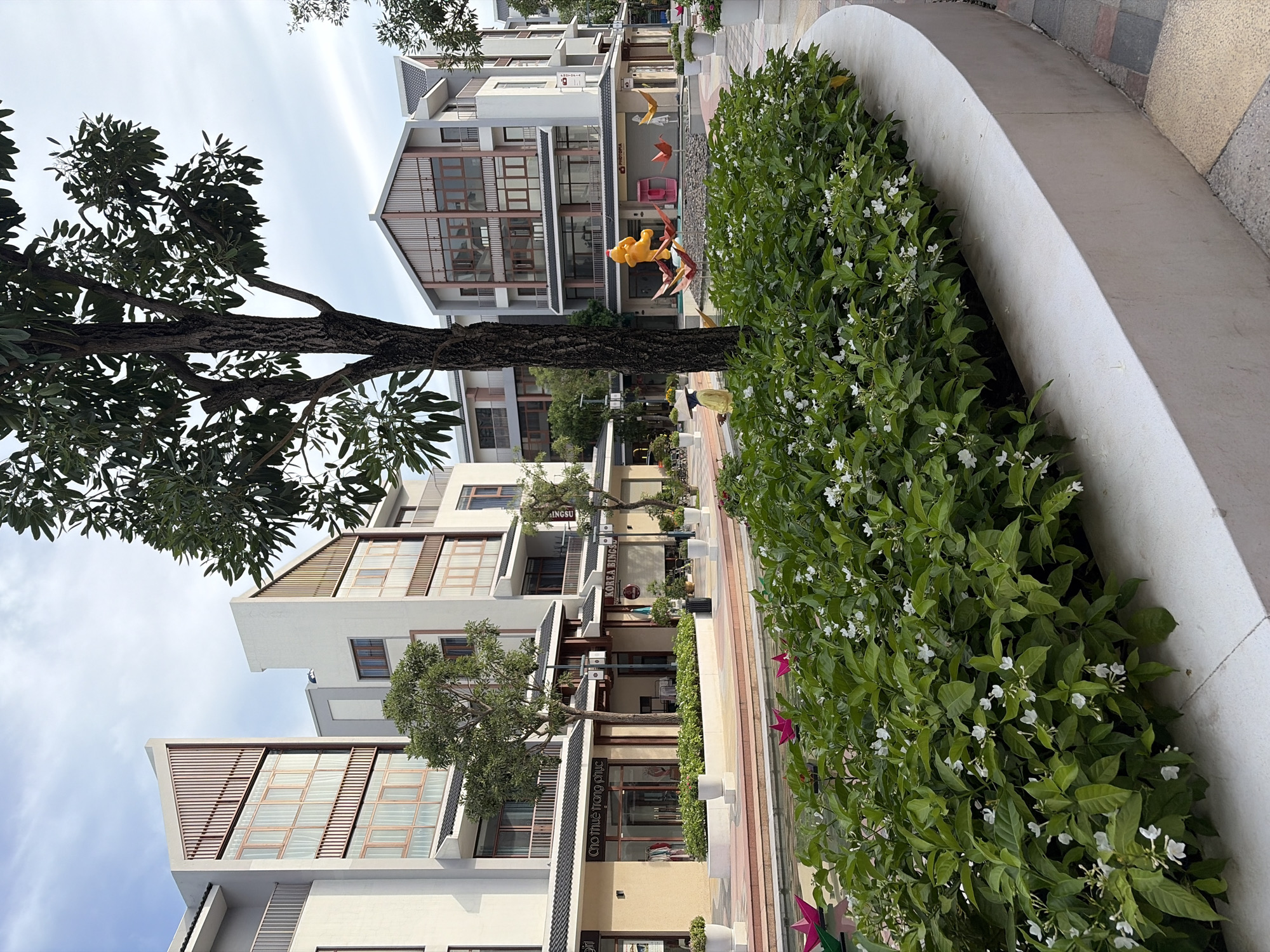 The image depicts a modern urban development in Nghĩa Trụ Commune, Vietnam, featuring a row of multi-story buildings with distinctive architectural elements. The buildings are white with grey accents and feature large windows with wooden frames, and some have angled, slatted wooden panels on their upper facades. In front of the buildings is a wide, paved walkway alongside a narrow canal or water feature.
A lush, manicured hedge of green bushes with small white flowers lines the edge of the walkway overlooking the water. The water feature contains several colorful, geometric origami sculptures, including pink flowers, yellow birds, and a large yellow cartoon character resembling Winnie the Pooh, as well as a pink sculptural element.
A large tree with a dark trunk and dense green foliage is prominently positioned in the foreground, partially obscuring the view of the buildings. The ground is paved with interlocking bricks in shades of red, grey, and yellow. A few white potted plants are visible along the walkway. In the middle ground, a person wearing a conical hat and a yellow outfit stands near the water feature. The sky is overcast with a few wispy clouds. The overall scene suggests a commercial or residential complex with a focus on aesthetic landscaping and public art.