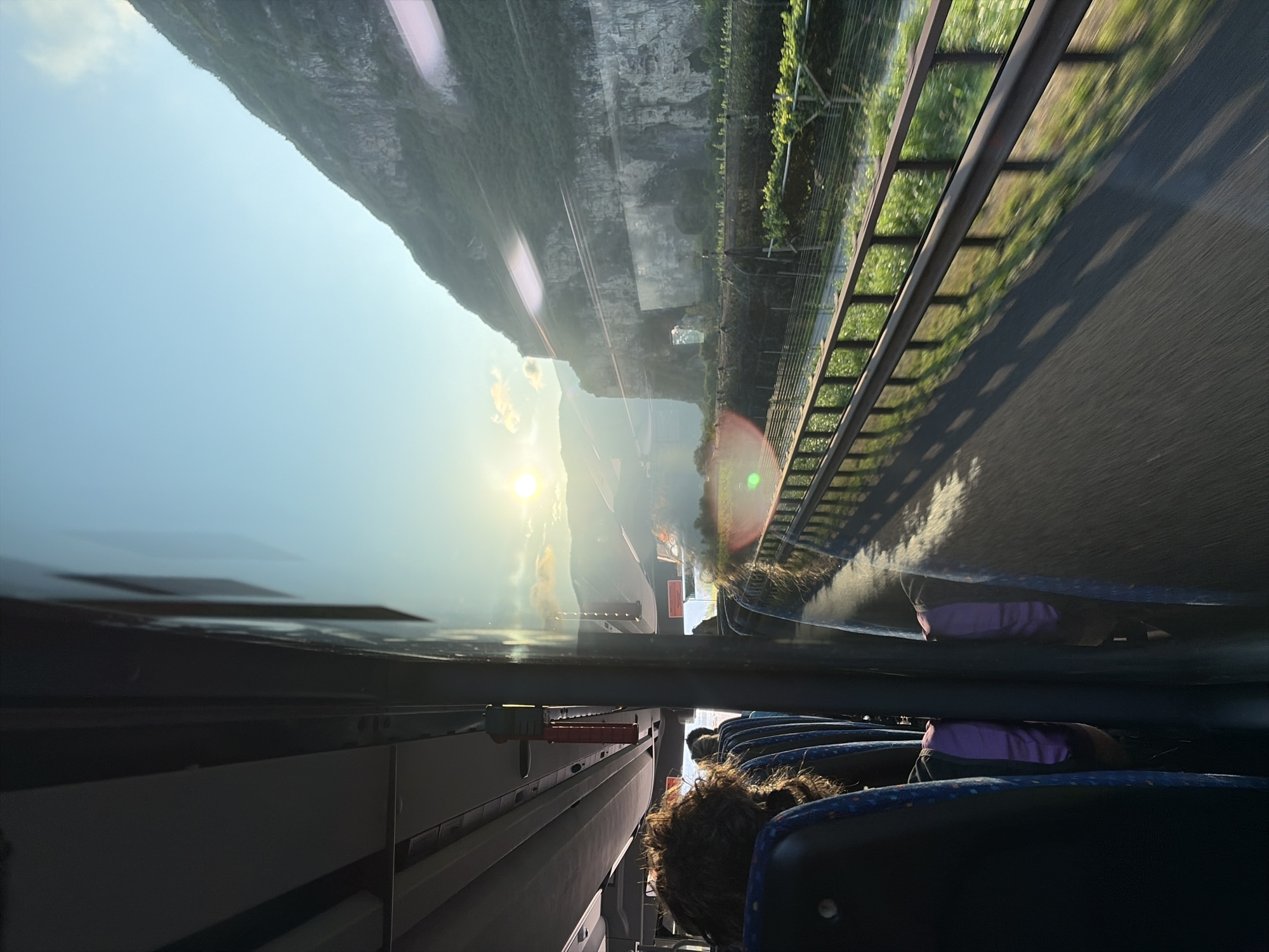 The image shows a view from inside a bus traveling on a road in Roveré della Luna, Italy. The sun is setting, casting a warm glow over the mountainous landscape. A steep, green-covered mountain dominates the right side of the frame, with a rocky cliff face visible. In the foreground, a metal guardrail runs alongside the road, bordered by lush green vegetation, likely vineyards. The road surface appears to be asphalt, showing motion blur indicative of the bus's movement. Inside the bus, passengers are visible, with the back of their heads and shoulders showing. One passenger in the foreground has curly brown hair. The seats are upholstered in blue fabric with a patterned design. The bus interior features overhead compartments and lighting fixtures. The scene is bathed in the golden light of the setting sun, creating long shadows on the road.