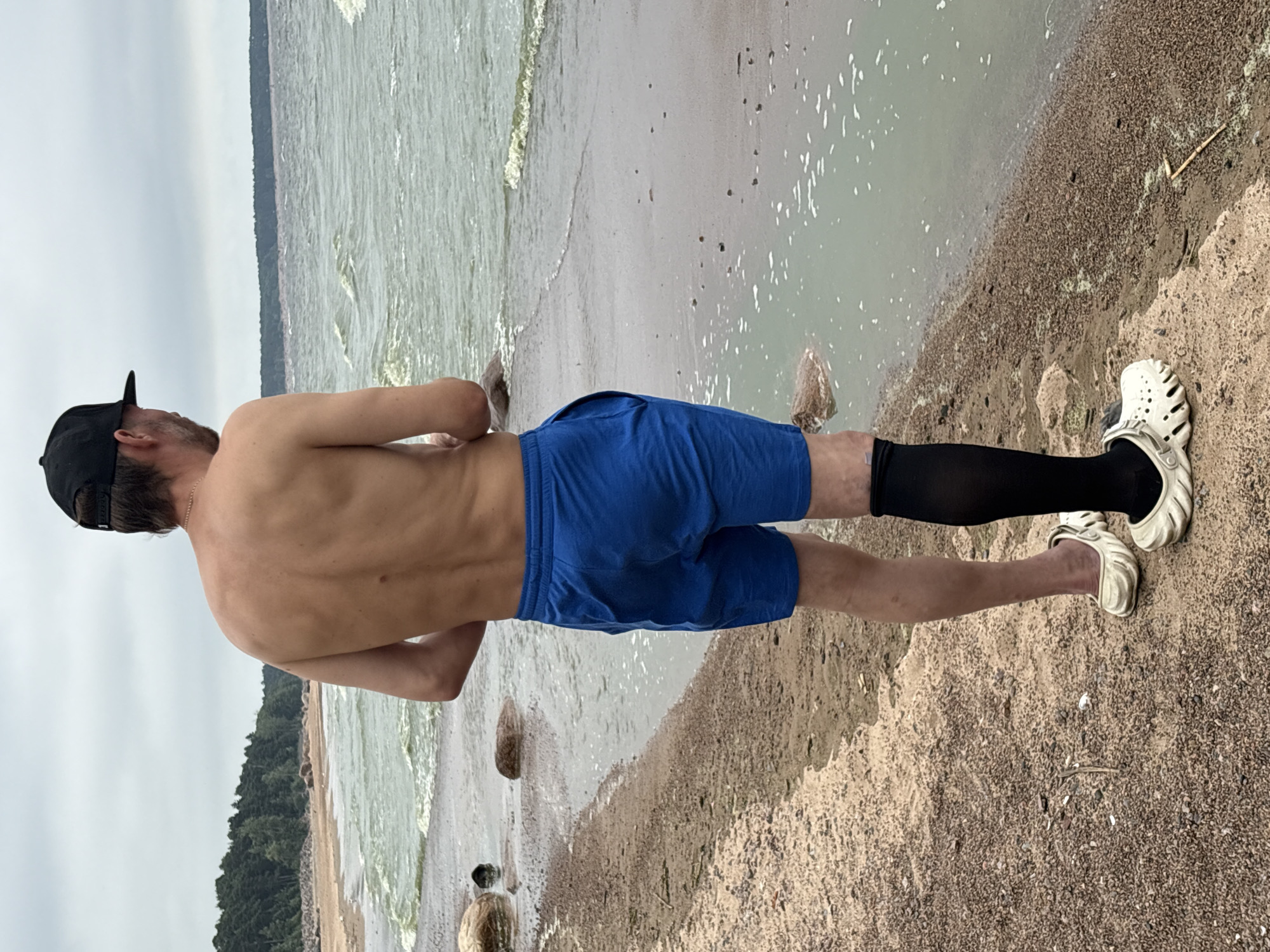 A bare-chested man wearing blue shorts, a black baseball cap, a black compression sleeve on his left leg, and white clogs stands on a sandy beach. He is facing away from the camera, looking out at a choppy sea under an overcast sky. The background features a line of trees and a distant horizon. The scene is located at Solnechnoye, Russia, on a beach with scattered rocks and some seaweed.