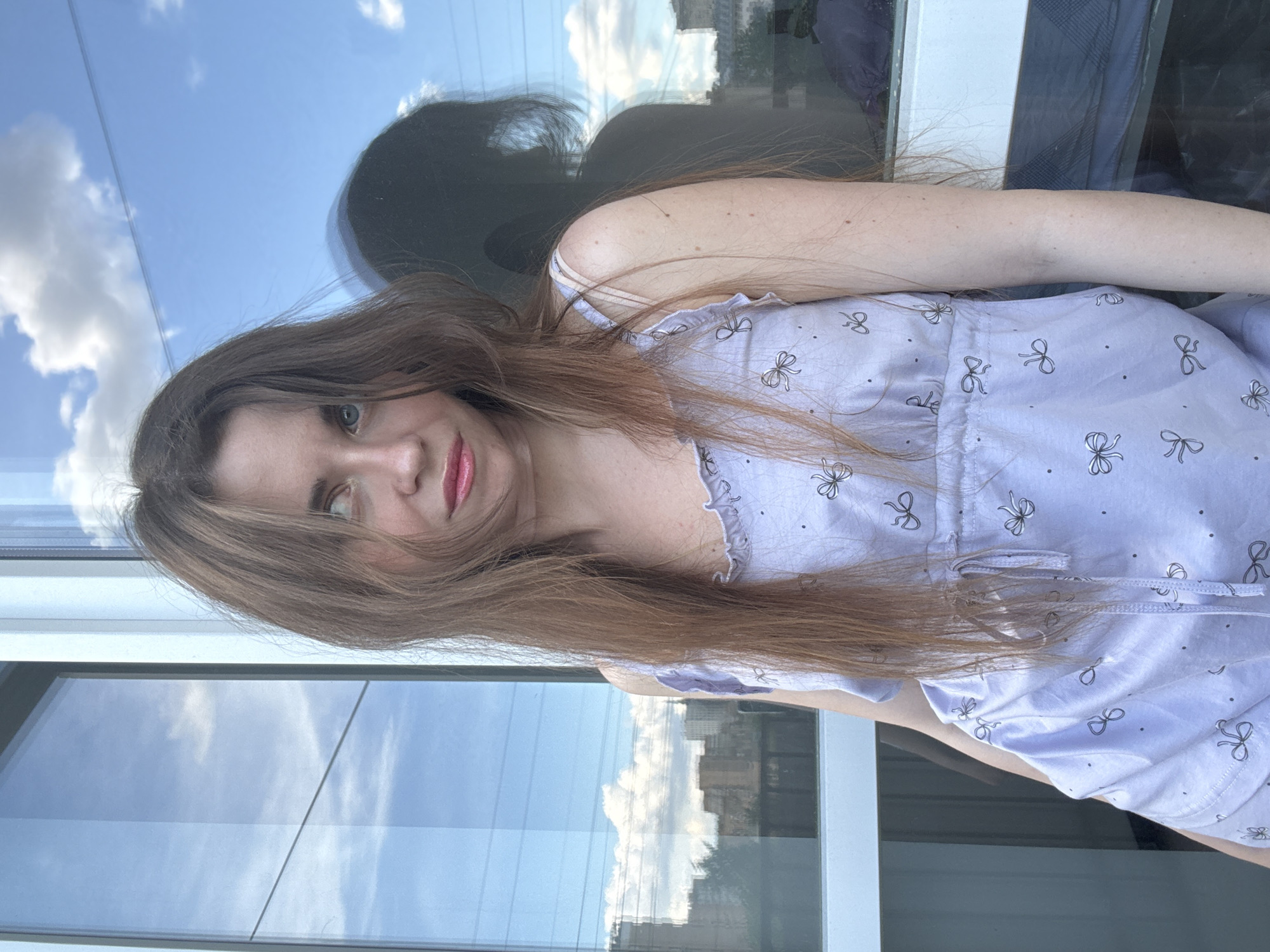 A young woman is facing the camera with her head tilted slightly. She has long, light brown hair that partially covers her left eye. She has blue eyes and pink lipstick. She is wearing a light purple dress with small bow patterns. Behind her is a window reflecting a blue sky with white clouds and the silhouette of a city, possibly Moscow, Russia. The window frame is visible on the sides.