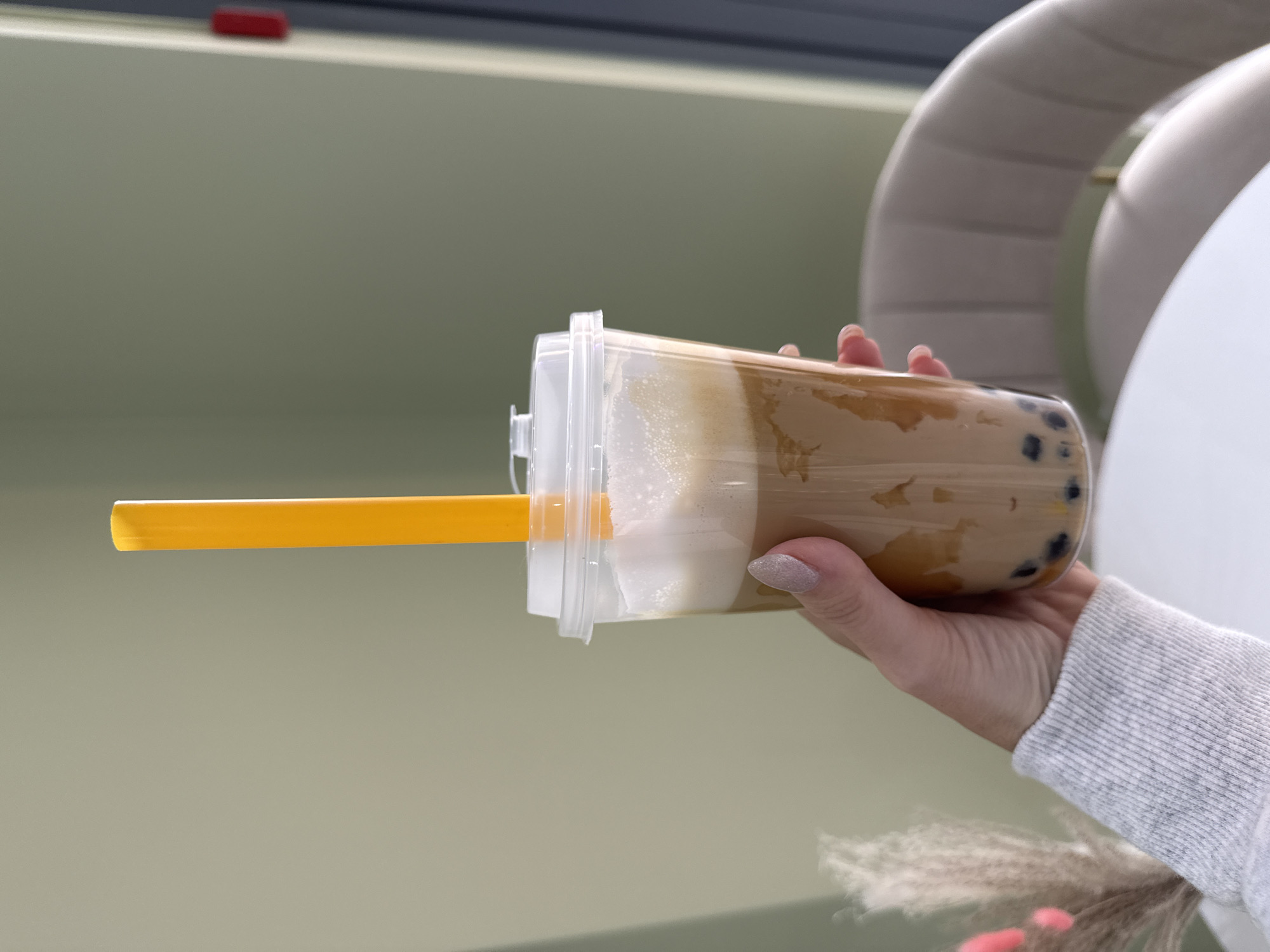 A person's hand, with a glittery manicure, holds a clear plastic cup filled with a layered beverage. The drink appears to be a brown, milky liquid with visible boba pearls at the bottom, topped with a layer of foam and a creamy white substance. A bright yellow, thick straw extends from the cup. The background is a soft, neutral green wall, with a hint of a curved, upholstered furniture piece in pale gray visible on the right. Above the green wall, a dark band with a small red rectangular object is seen, possibly part of a shelf or fixture. The lighting suggests it is daytime, likely indoors in a well-lit space. The scene is focused on the beverage and the hand holding it, implying a casual moment of enjoyment or preparation of the drink. No text is visible in the image. The context of Moscow, Russia is noted but not explicitly depicted within the visual elements of the image itself.