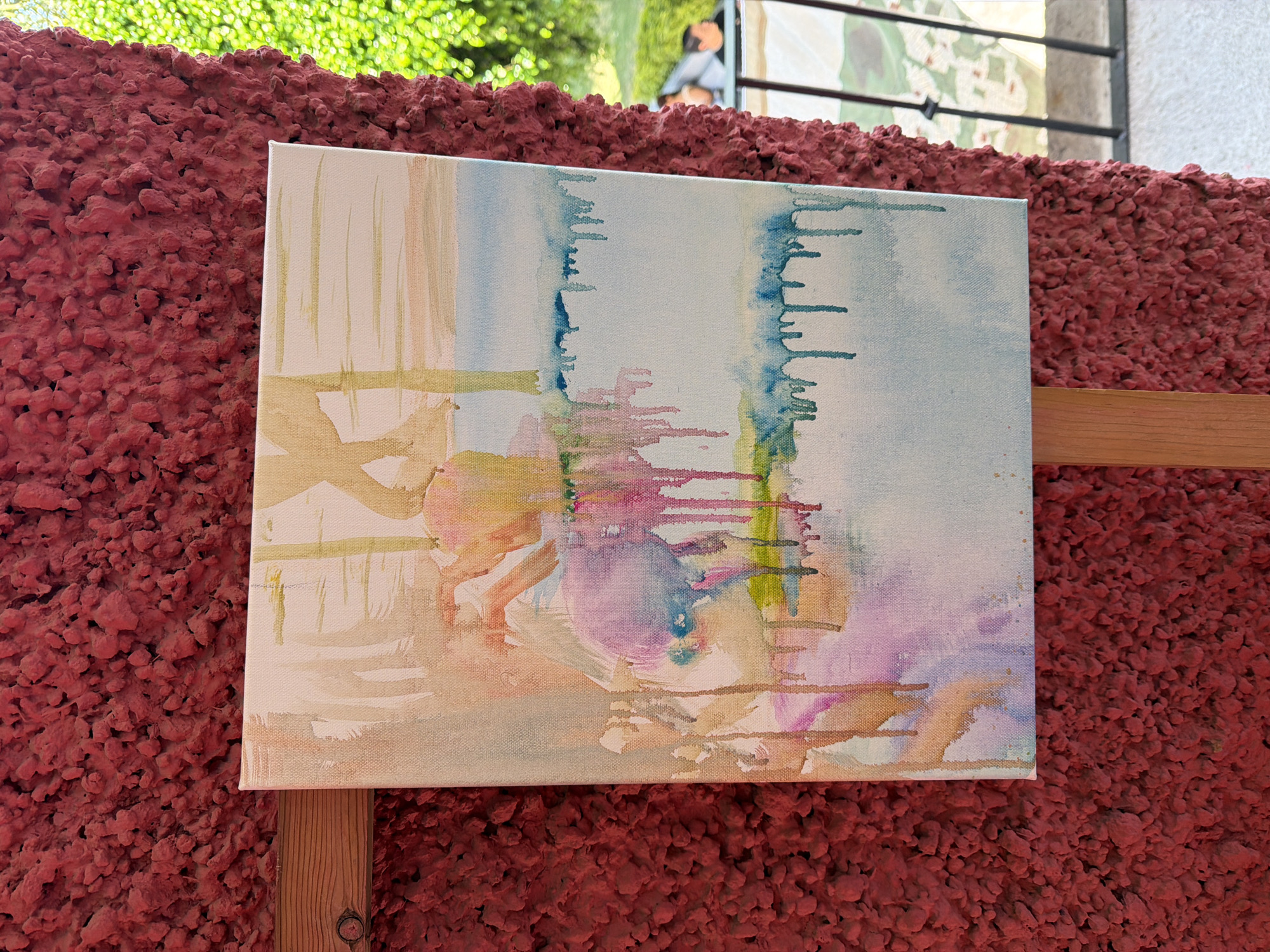 A rectangular canvas painting is displayed outdoors in San Andrés Cholula, Mexico. The painting, an abstract watercolor, features pale blues, pinks, yellows, and light browns with a vertical orientation and a dripping effect. The canvas is resting on a wooden easel. Behind the painting, there is a textured, dark-red wall. Green foliage is visible above the wall, partially obscuring a structure with a person. A portion of the building wall and frame can be seen to the right of the foliage, over the painting.