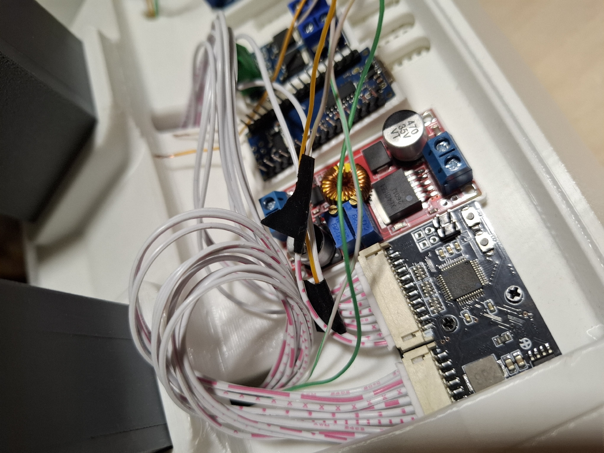 The image shows a close-up view of electronic components and wiring within a white plastic enclosure. Several circuit boards, including what appears to be a microcontroller board with a large chip and several smaller surface-mount components, are visible. There are also other smaller blue circuit boards with what look like voltage regulators or stepper motor drivers. A bundle of multi-colored wires, mostly white with pink and red stripes, is coiled in the upper left of the image and connects to one of the circuit boards. Other individual wires in green, yellow, and orange are also present, connecting various components. A toroidal inductor, a capacitor labeled "470 35V", and screw terminals are also visible on one of the circuit boards. The scene is illuminated from above, casting subtle shadows. The background consists of the white interior of the enclosure and parts of a dark gray object, possibly a larger enclosure or housing. There are no people, animals, or indications of the city of Tyumen, Russia.