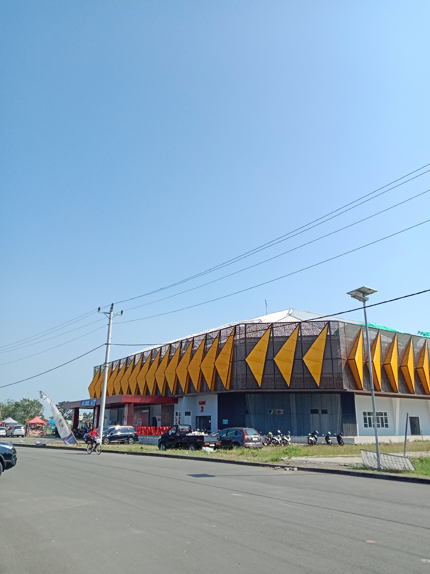 This is a medium-distance, eye-level shot of a modern building with a distinctive yellow and patterned facade, situated along a street in Kendal, Indonesia. The building appears to be a commercial establishment, possibly a market or retail center, with signs indicating "GATES 2."
The sky is a clear, bright blue with a few thin, dark power lines stretching across the upper portion of the frame. A utility pole with insulators stands to the left of the building, also supporting these power lines. To the right of the building, a solar-powered street lamp is visible.
In the foreground, a paved road runs horizontally across the image. To the left of the road, near the building's entrance, a person is riding a bicycle and carrying a large, tattered white flag. Several parked cars and motorcycles are visible in front of and to the side of the building, suggesting ongoing activity. A tent and some market stalls are present further to the left, indicating a potential market or roadside vendors. The ground beside the road is covered in grass and some patches of dirt.
The building itself has a white lower section with windows and doors, and an upper section characterized by a dynamic, angular design featuring alternating yellow triangular panels and a dark, patterned screen. The overall impression is one of a sunny day in an urban or semi-urban setting in Kendal.