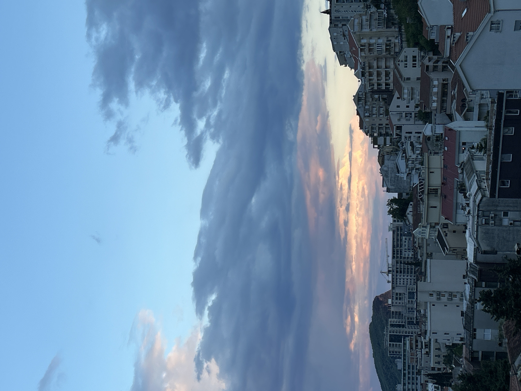 The image presents a panoramic view of an urban landscape under a dramatic sky, likely at dusk or dawn. The sky occupies the upper portion of the frame, transitioning from a clear, light blue on the left to an expanse of dark, heavy clouds in the center. Towards the right, these clouds are illuminated with warm pink, orange, and golden hues, suggesting the light of a setting or rising sun breaking through.
Below the expressive sky lies a dense cluster of multi-story buildings, characteristic of a coastal town. These structures, visible across the middle and lower right of the image, feature varied architectural styles, numerous balconies, and many reddish-brown tiled roofs. They appear closely packed, extending along what seems to be a sloping terrain. In the background, behind some of the buildings on the right, a green, tree-covered mountain or large hill is visible.
No people or explicit activities are depicted, focusing instead on the interplay between the urban environment and the natural elements of the sky and mountain. The scene, consistent with the provided location context of Budva, Montenegro, captures the town's typical blend of dense development against a backdrop of mountainous coastal scenery. There is no visible text in the image.