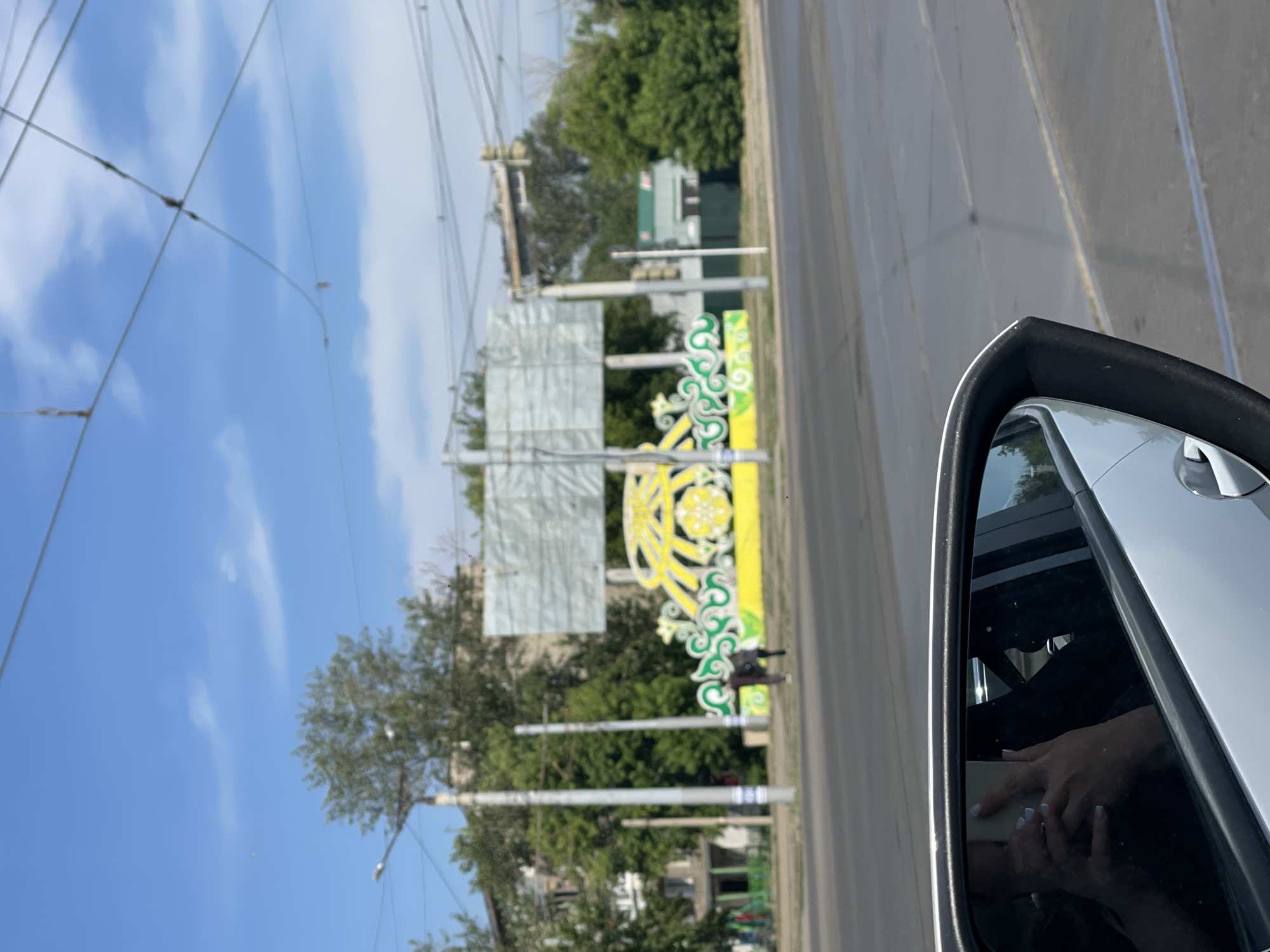 A reflection of a person holding a smartphone is visible in the side mirror of a white car. The car is on a street in Pavlodar, Kazakhstan, with a view of a public area with trees, buildings, and power lines. In the background, there is a large, ornate yellow and green structure with a billboard behind it. Two figures, likely pedestrians, are walking near the structure. The sky is blue with some scattered clouds.