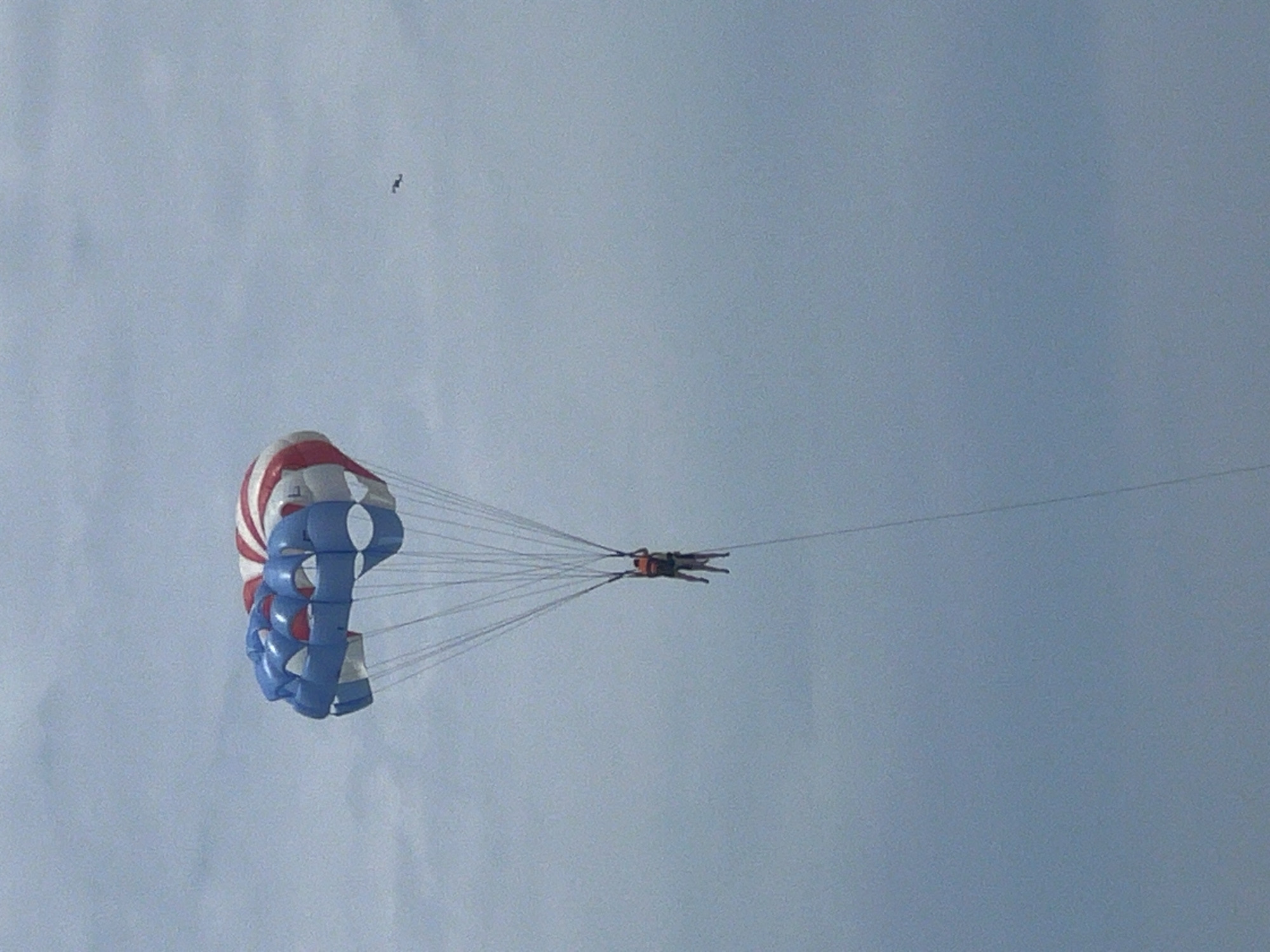 A parasail with a red, white, and blue striped canopy is airborne against a cloudy, pale blue sky. Two individuals are suspended beneath the canopy, attached by harnesses and ropes. The person in front wears an orange life vest. A second, smaller silhouette of what appears to be a bird or kite is visible in the upper right quadrant of the sky, distant from the parasail. The image was captured from a low angle looking upwards. This scene is likely taking place off the coast of Đà Nẵng, Vietnam, a popular tourist destination known for its beaches and water activities.