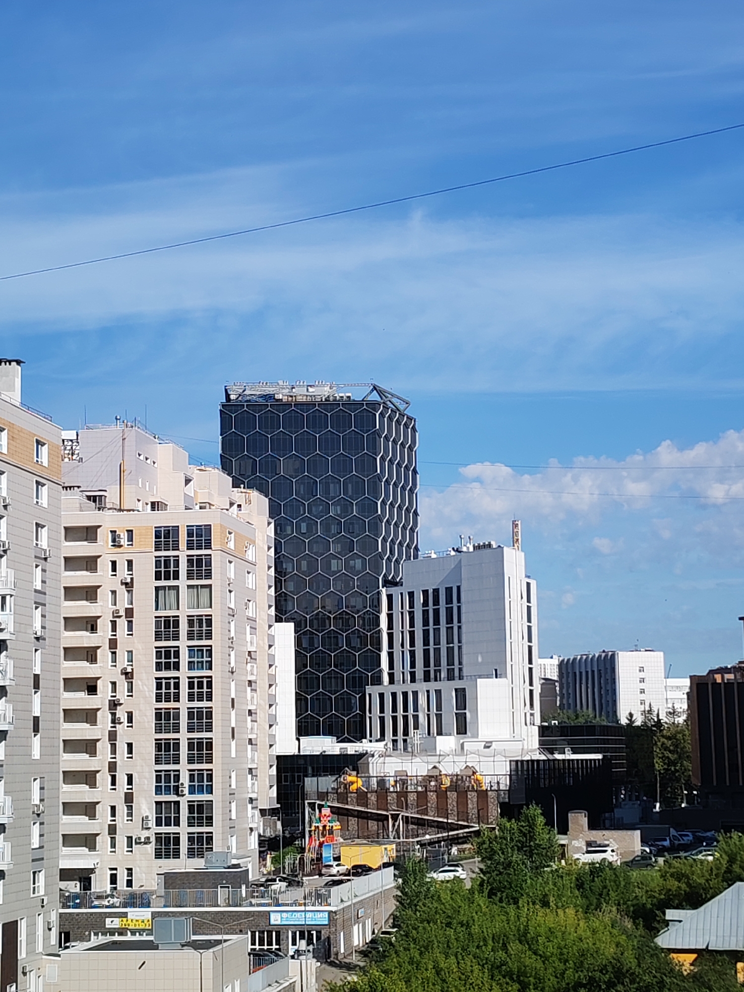 The image depicts a cityscape in Ufa, Russia, on a clear day with a blue sky dotted with wispy clouds. The foreground features several modern apartment buildings with light-colored facades and numerous windows and balconies. To the right and further back, a striking, tall skyscraper with a distinctive hexagonal pattern on its dark facade stands prominently. Adjacent to it is a white, multi-story building with a modern, angular design, and further to the right, other office buildings and some construction activity are visible. The scene includes a wire running diagonally across the upper part of the image, likely a power line. The lower part of the image is partially obscured by lush green trees. There are no people visible in the image.
