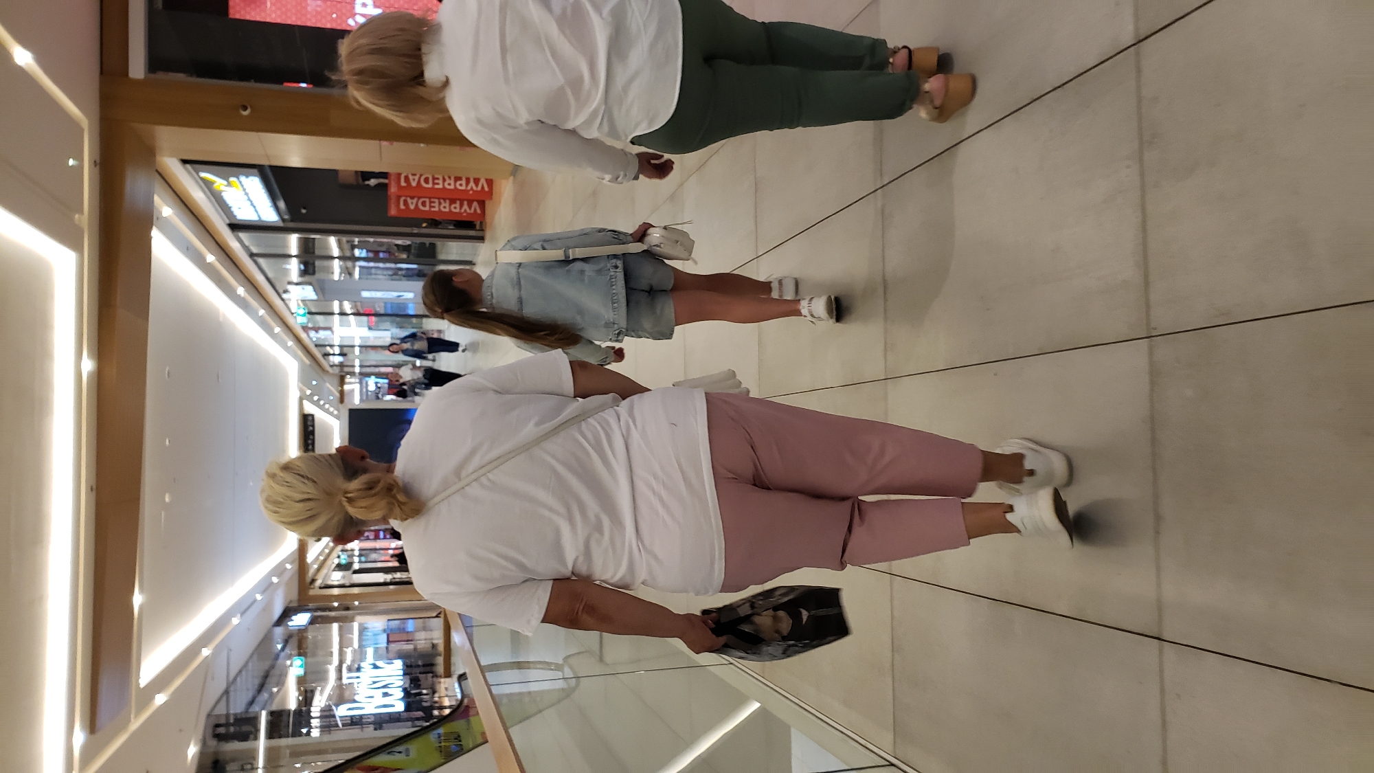 This image depicts an interior scene, likely a shopping mall, with a group of people walking down a tiled corridor. The lighting suggests an indoor environment, possibly a commercial establishment.
In the foreground, a woman with blonde hair tied in a bun is walking away from the camera. She is wearing a white t-shirt and pink trousers. She is carrying a black shopping bag in her left hand. To her right, a young girl with long brown hair in a ponytail is walking ahead, wearing a light blue denim jacket and denim shorts. She has a white crossbody bag. Further to the right, another woman with blonde hair is walking away from the camera, wearing a white shirt and green trousers. She is wearing platform sandals.
The corridor features a polished tiled floor with dark grout lines. The ceiling is illuminated with strip lighting integrated into a suspended structure. Storefronts with glass windows are visible on both sides of the corridor, with signage indicating retail establishments. One sign visible in the background on the left reads "Bershka". On the right, signs with "VÝPREDAJ" (Slovak for "SALE") are displayed. The architecture suggests a modern shopping center.
The context of Prešov, Slovakia, is provided for the location. This image captures a common scene in a modern shopping environment, focusing on the movement and attire of individuals within this public space.