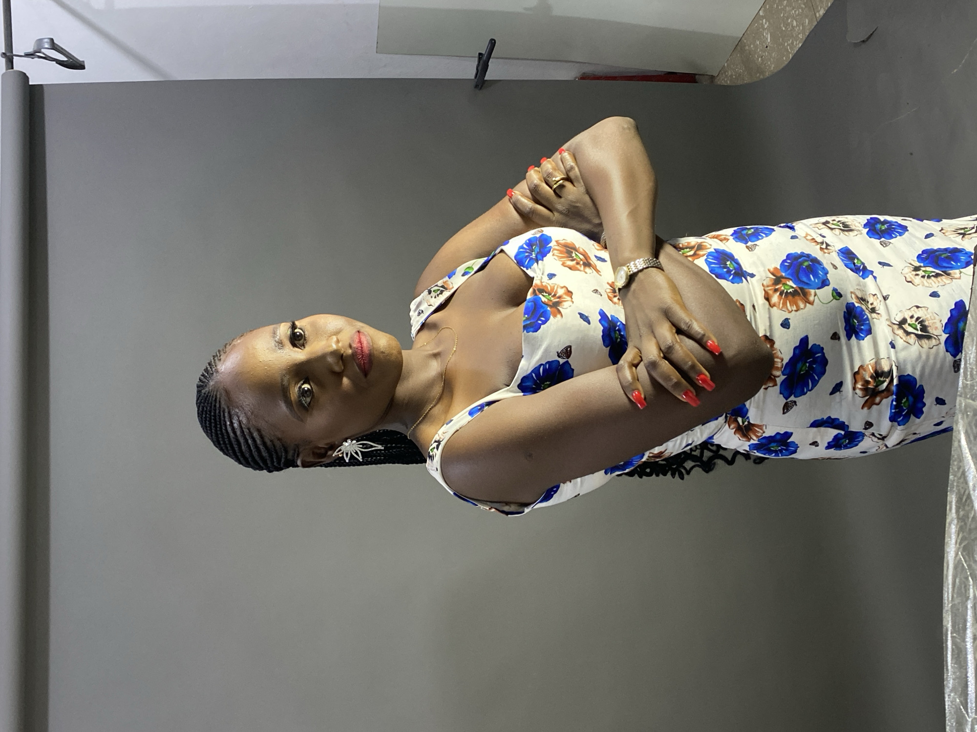 A woman is featured in a full-body portrait, standing against a plain grey studio backdrop, suggestive of an indoor photography session. She looks directly at the camera with a composed expression, her head slightly tilted, and her lips accented with red lipstick. Her arms are crossed in front of her chest.
She is wearing a white sleeveless dress patterned with prominent blue and earthy-toned floral designs. Her dark hair is styled in neat braids, revealing an intricate star-shaped earring on her left ear. Her fingernails are painted a striking red, complementing her lipstick. A silver-toned watch adorns her left wrist, and a ring is visible on her right hand. A thin, gold-colored necklace is also visible around her neck.
The background shows a large roll of grey backdrop paper on the left side of the frame and what appears to be studio equipment, possibly a light stand or backdrop support, near the top left corner. The lighting is even, highlighting the subject clearly. This image captures a posed photoshoot, with the location context being Jos, Nigeria. No text is visible in the image.