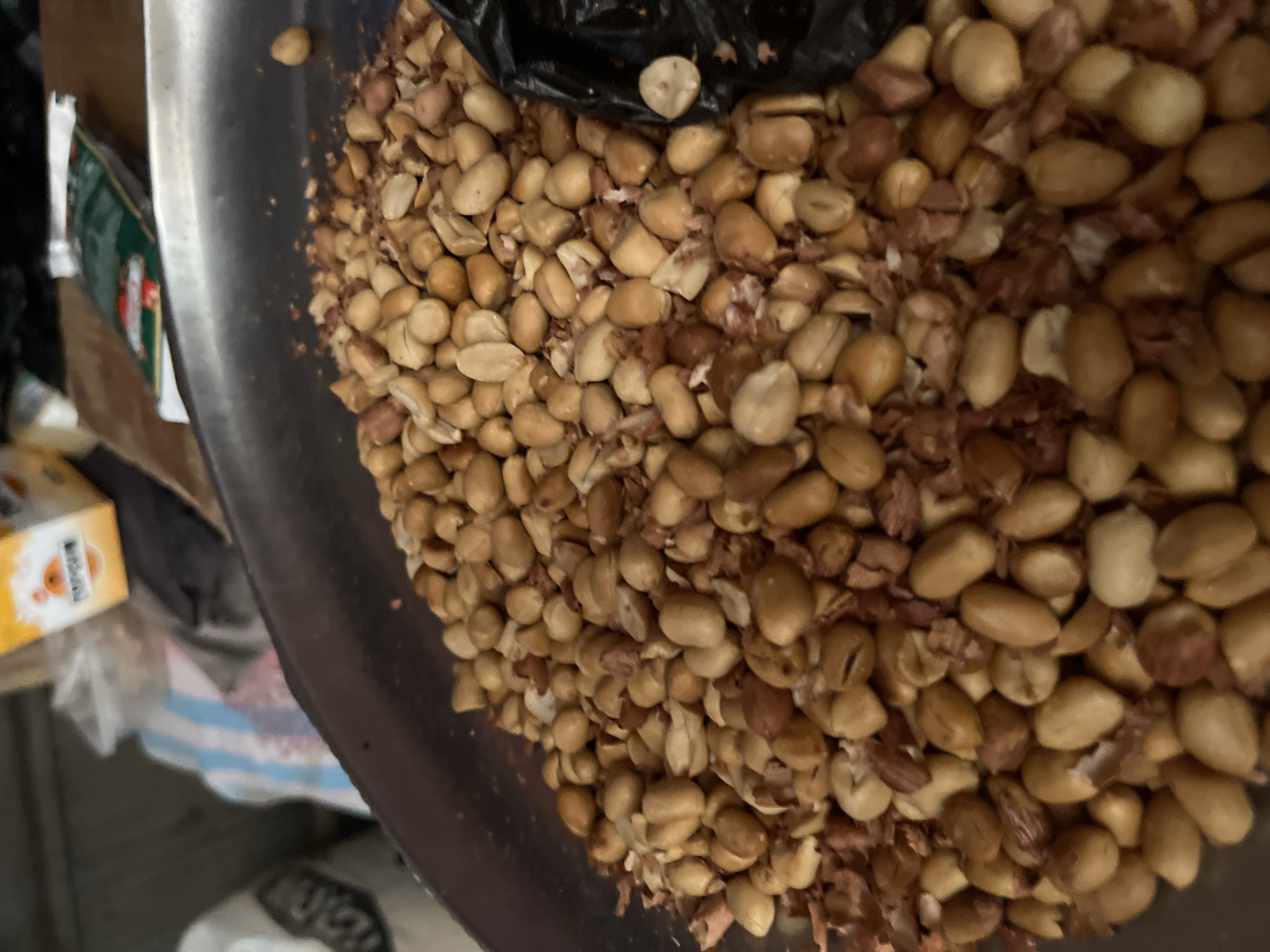 A close-up, overhead shot shows a pile of shelled peanuts, some halved, in a large, shallow metal bowl. The peanuts are a light tan color, with some showing reddish-brown skins. The bowl appears to be on a table. In the background, a blurry yellow and orange box with writing is visible, along with what appears to be a plastic bag and some striped fabric. To the right of the bowl, a portion of a black plastic bag is visible, filled with peanuts. The scene is indoors, likely in a market or food stall, given the context of produce being sold. The image does not provide specific information about the city of Abuja, Nigeria, other than it being a potential location for such a scene.