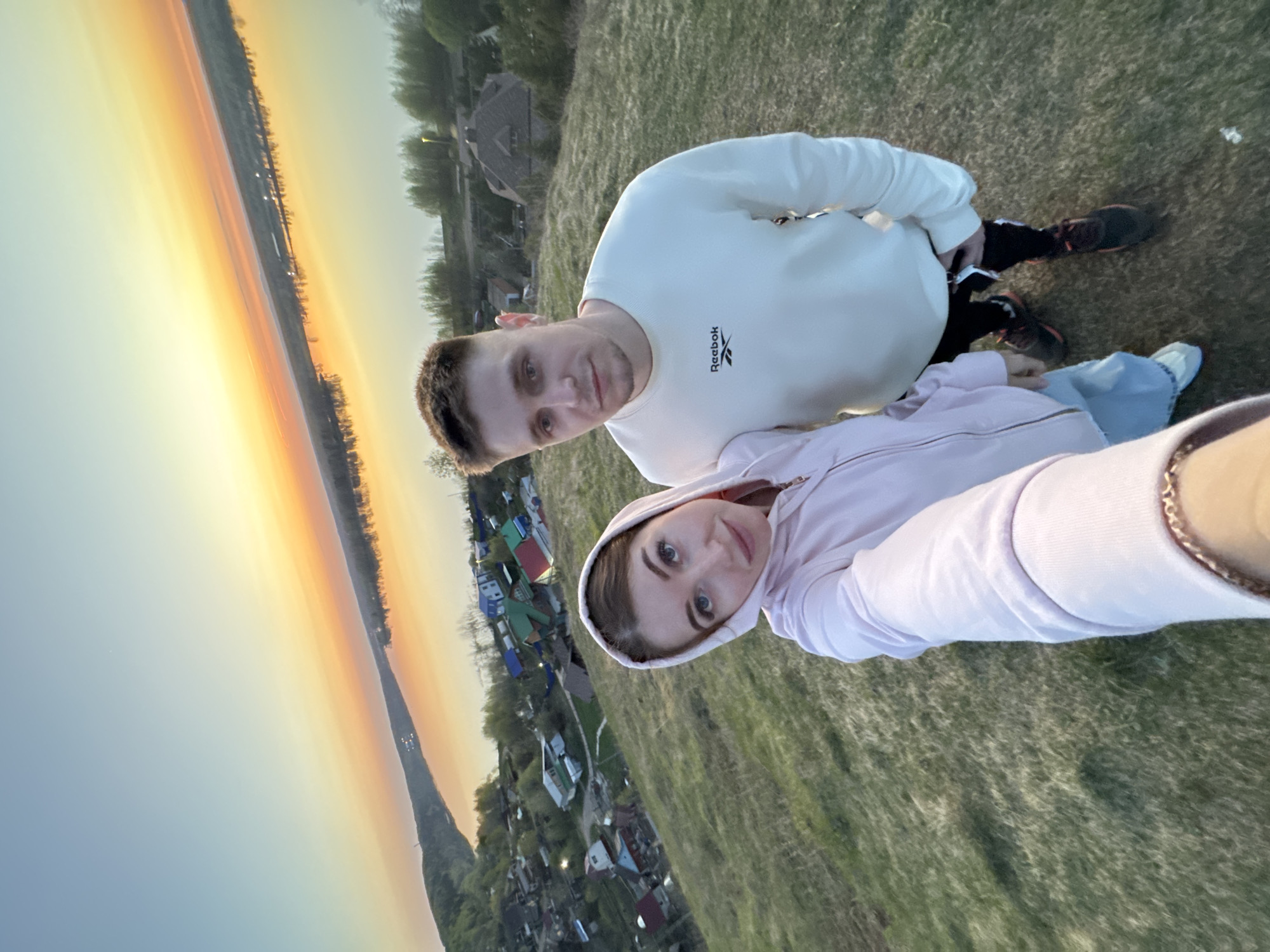 A selfie of a fair-skinned woman in a pink hooded sweatshirt and a fair-skinned man in a white Reebok sweatshirt, taken outdoors at dusk, possibly in the city of Ольховка, Russia. The couple are lying on grassy ground in front of a landscape that includes a body of water reflecting the sunset, and a village with houses and trees. The woman is holding the camera, and both people are looking towards the lens. The man is positioned slightly behind and above the woman.