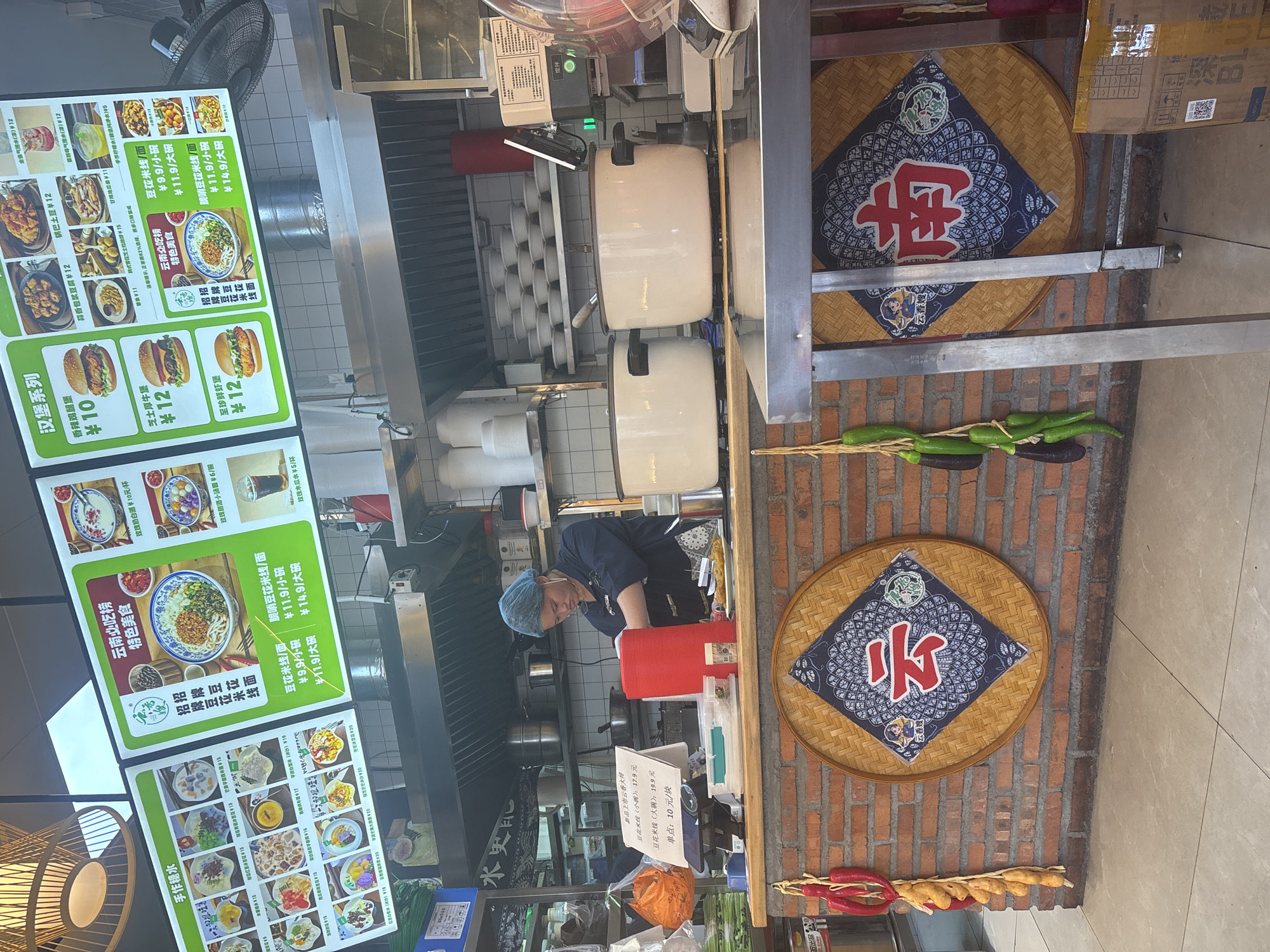 This image depicts a fast-food restaurant counter area in Suzhou Industrial Park, China. The scene is focused on a food display and preparation zone, with a worker in the background.
On the left, a bright green menu board is prominently displayed. It features various food items with accompanying images and prices in Chinese characters. Some of the visible categories include "手作糖水" (Handmade Sweet Soups) and "汉堡系列" (Hamburger Series). Another menu board to the right, also green, highlights "云南南烤" (Yunnan Southern Roast) and "特色美食" (Specialty Foods), with a large image of a noodle dish and its price.
In the background, behind the counter, a person wearing a blue hairnet and a dark uniform is visible, appearing to be involved in food preparation. They are wearing a face mask. Behind the worker, shelves are stocked with bowls, and kitchen equipment like pots and a ventilation hood are present.
The counter itself is made of brick and wood. On the counter, there are several food items, containers, and a cash register. A large red cylindrical object is to the right of the worker.
Decorations in the scene include two large round woven mats on the brick wall, each displaying a traditional Chinese character in the center: "云" (Yun) on the left and "南" (Nan) on the right. Bunches of red chilies and what appear to be small dried gourds or sausages hang on the left side of the frame.
The overall environment suggests a busy, casual dining establishment. The lighting appears to be a mix of overhead artificial lights and natural light.