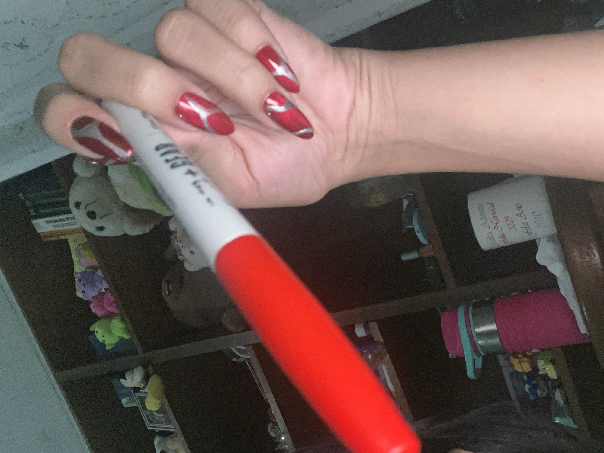 The image depicts a close-up view of a hand with red, almond-shaped nails adorned with silver accents, holding a large red and white marker. The marker is positioned diagonally, with its red end extending towards the bottom left and its white end towards the top right. The background consists of a bookshelf filled with various stuffed animals and other items, suggesting an indoor setting. The lighting is dim, and there is a slight blur throughout the image. The location of Santa Ana, Venezuela is not discernible from the provided image.