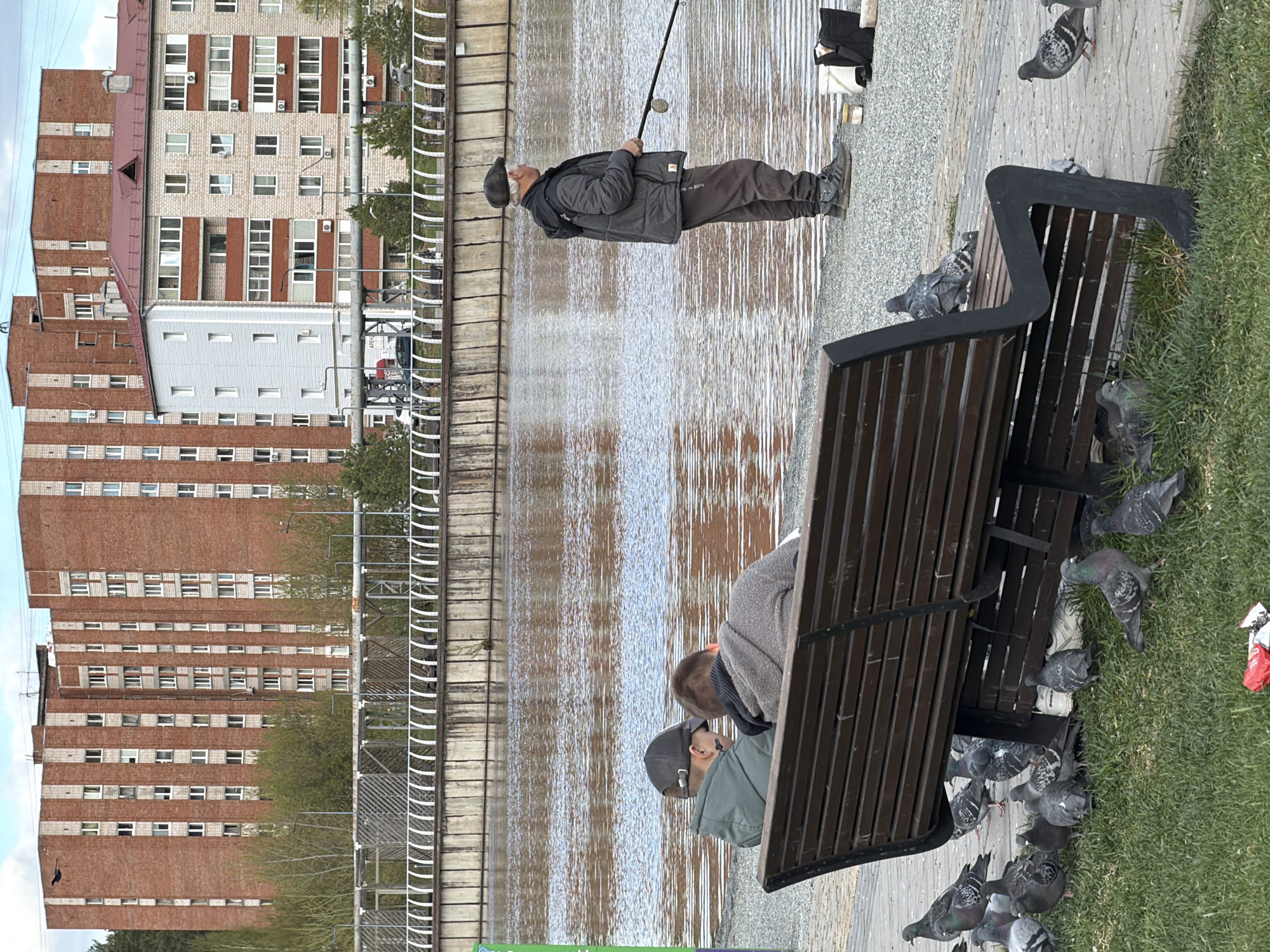 An eye-level, outdoor shot of a canal in Votkinsk, Russia, with people and pigeons. The canal runs horizontally across the center. A man with a fishing pole, wearing a dark-colored hat, a black jacket, and brown pants, stands along the water’s edge. Two men are sitting on a bench on the right side of the photo. Pigeons are on the bench and the grass near the bench. In the background, a tall brick building with light-colored windows is visible on the other side of the canal. A gray metal fence is along the edge of the water. The sky is blue with some clouds.