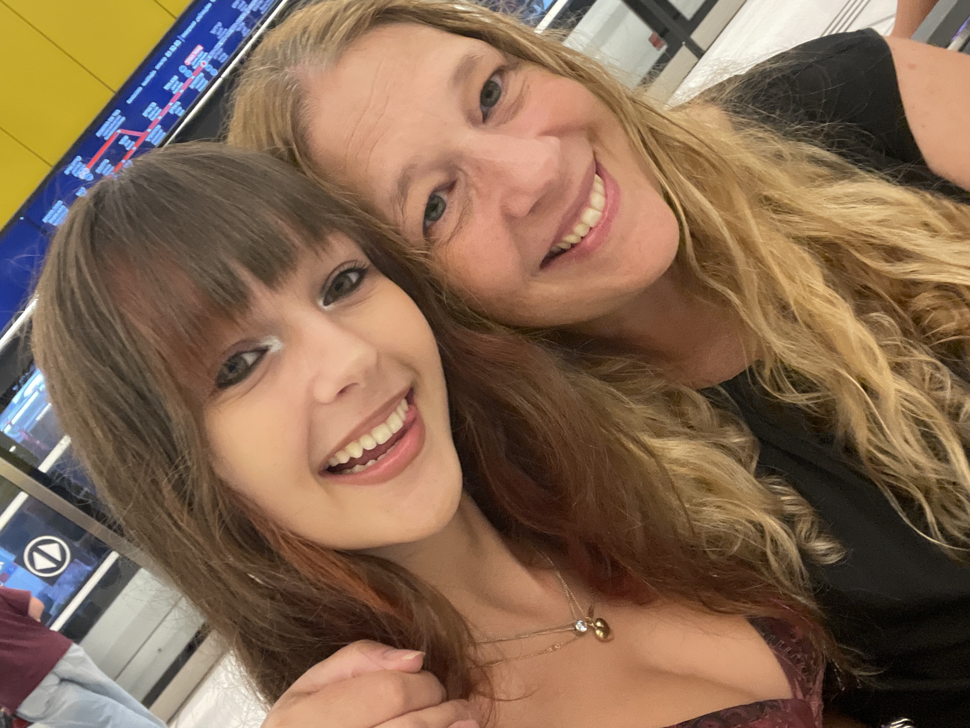 A selfie taken in a well-lit, modern indoor location, likely a public transportation hub, is presented. The foreground is dominated by the faces of two individuals. On the left, a younger person with long, brown hair, bangs, and a slight smile is positioned closer to the camera, their head tilted. They are wearing a dark-colored garment with a visible necklace featuring a small, heart-shaped pendant. To their right and slightly behind, an older person with blonde hair and a warm smile is also looking towards the camera. They are wearing a black shirt. The background shows a yellow wall with a dark blue digital display board showing what appears to be route information or a timetable, a common sight in Tel-Aviv's train stations or bus terminals. To the far left, a portion of another person wearing a maroon shirt and light blue pants is visible, facing away from the camera, suggesting they are waiting for transportation. The overall scene conveys a casual and positive moment captured during a journey.