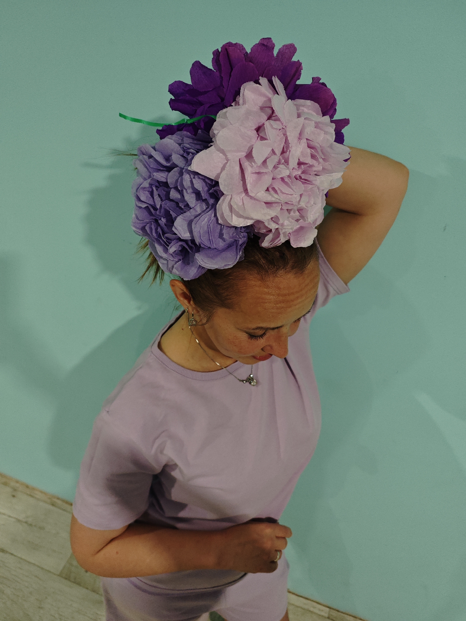 A high-angle shot shows a woman with light skin and freckles standing indoors against a turquoise wall. She wears a light purple t-shirt and shorts, also light purple, appearing to be a matching set. Her right arm is raised and bent at the elbow, the hand positioned behind her head.
A bouquet of three large, paper flowers in shades of purple and pink sits atop her head. A green stem is visible amongst the flowers. She wears dangling earrings and a necklace with a small pendant. Her head is slightly bowed, and she looks downward. Her left hand is near her stomach. The location, Markovo, Russia, is not identifiable from the image.