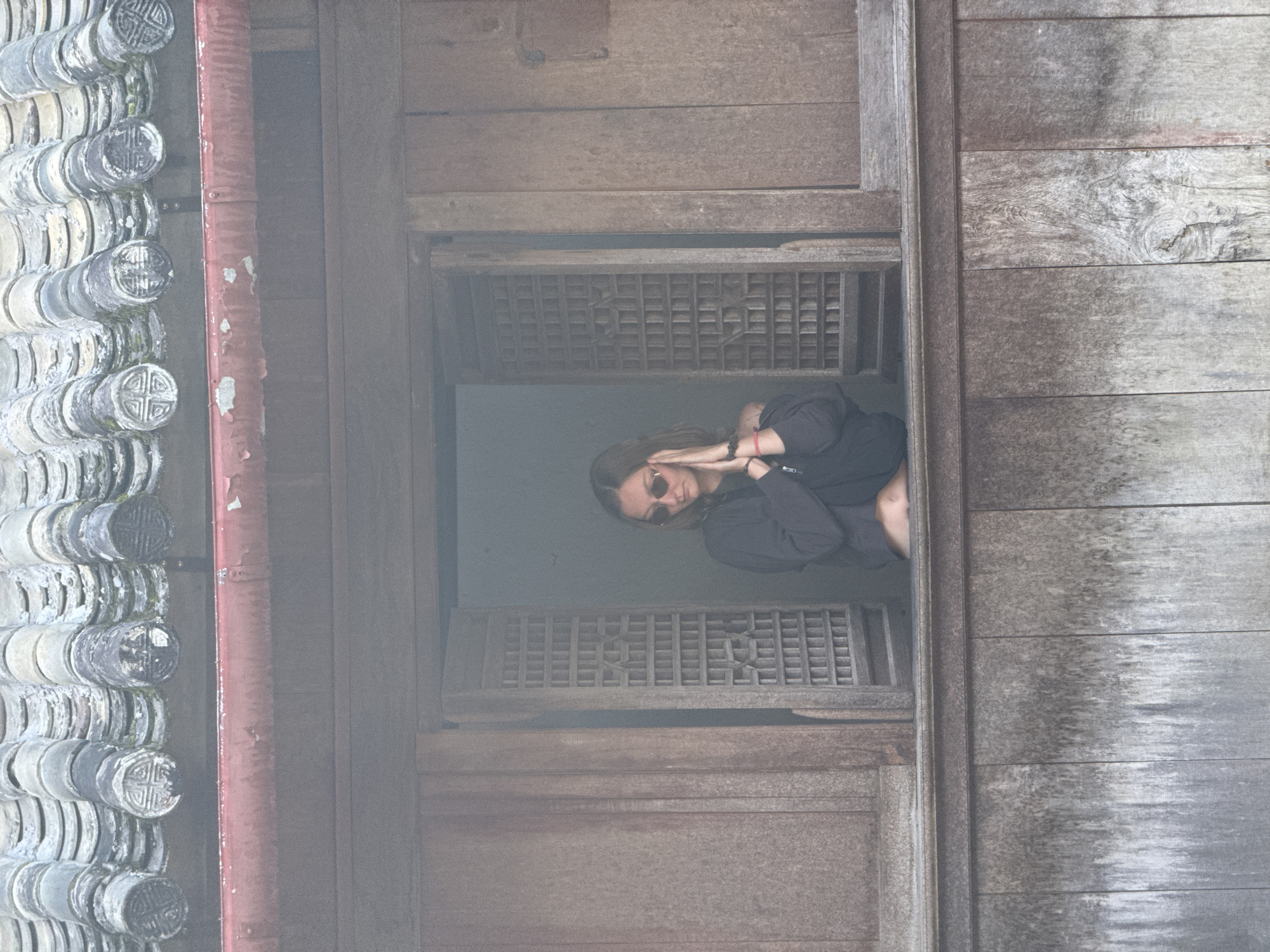 A woman wearing sunglasses and a dark long-sleeved shirt looks out of a window with a lattice grill. The structure of the window and surrounding walls is wooden. Old, patterned, ceramic roof tiles line the edge of the frame to the left. The scene is in Sà Phìn Commune, Vietnam.